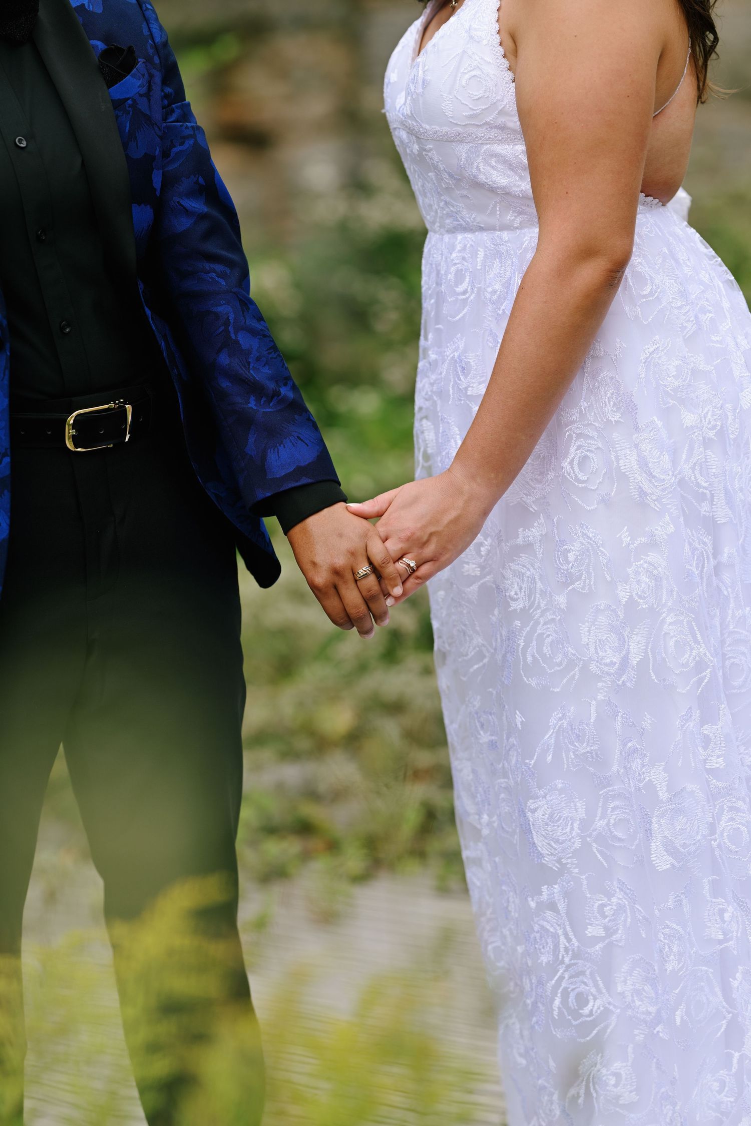 Close-up of bride and groom holding hands, bride in white lace gown, groom in blue jacket.