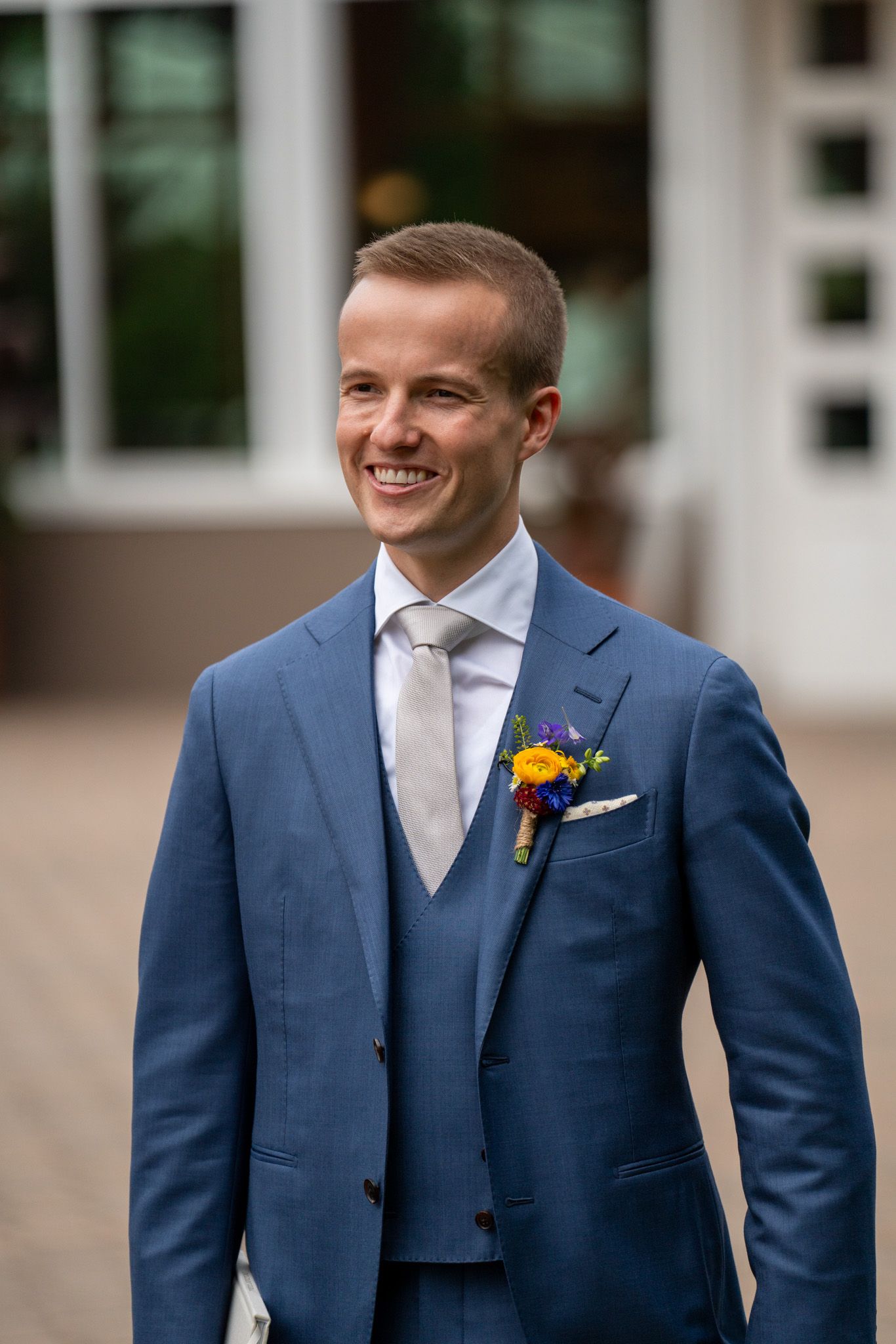 A groom in a tailored blue three-piece suit with yellow boutonniere smiles during his wedding day.