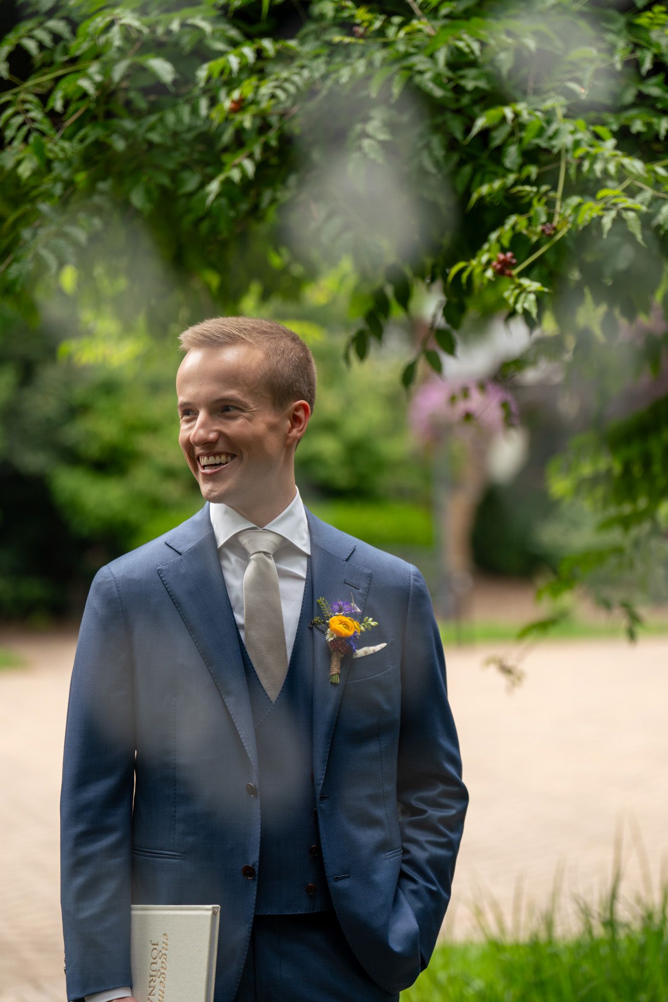 A groom stands outdoors in a navy suit holding a book surrounded by green foliage.