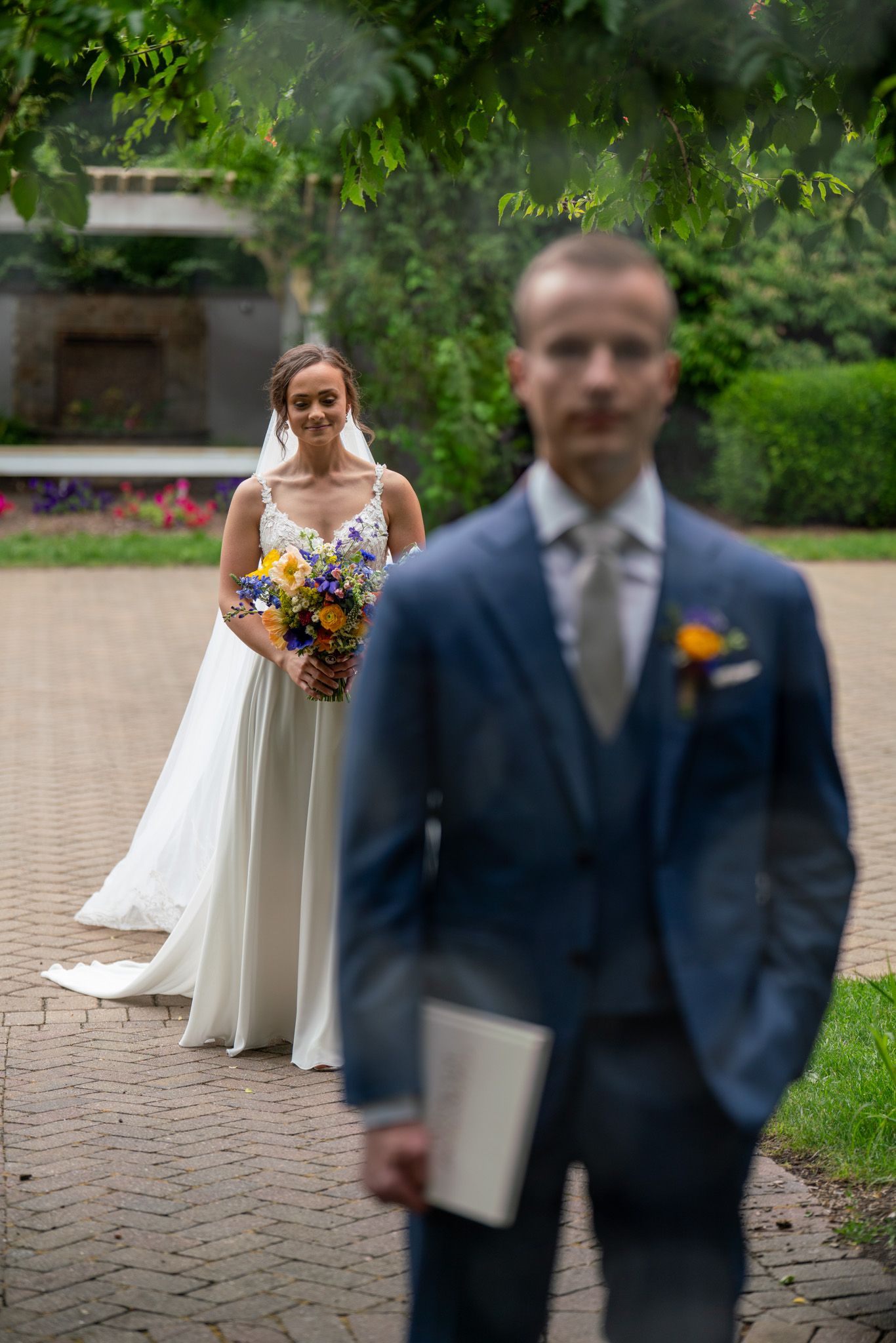 A wedding first look moment with bride in white gown holding flowers behind groom in blue suit.