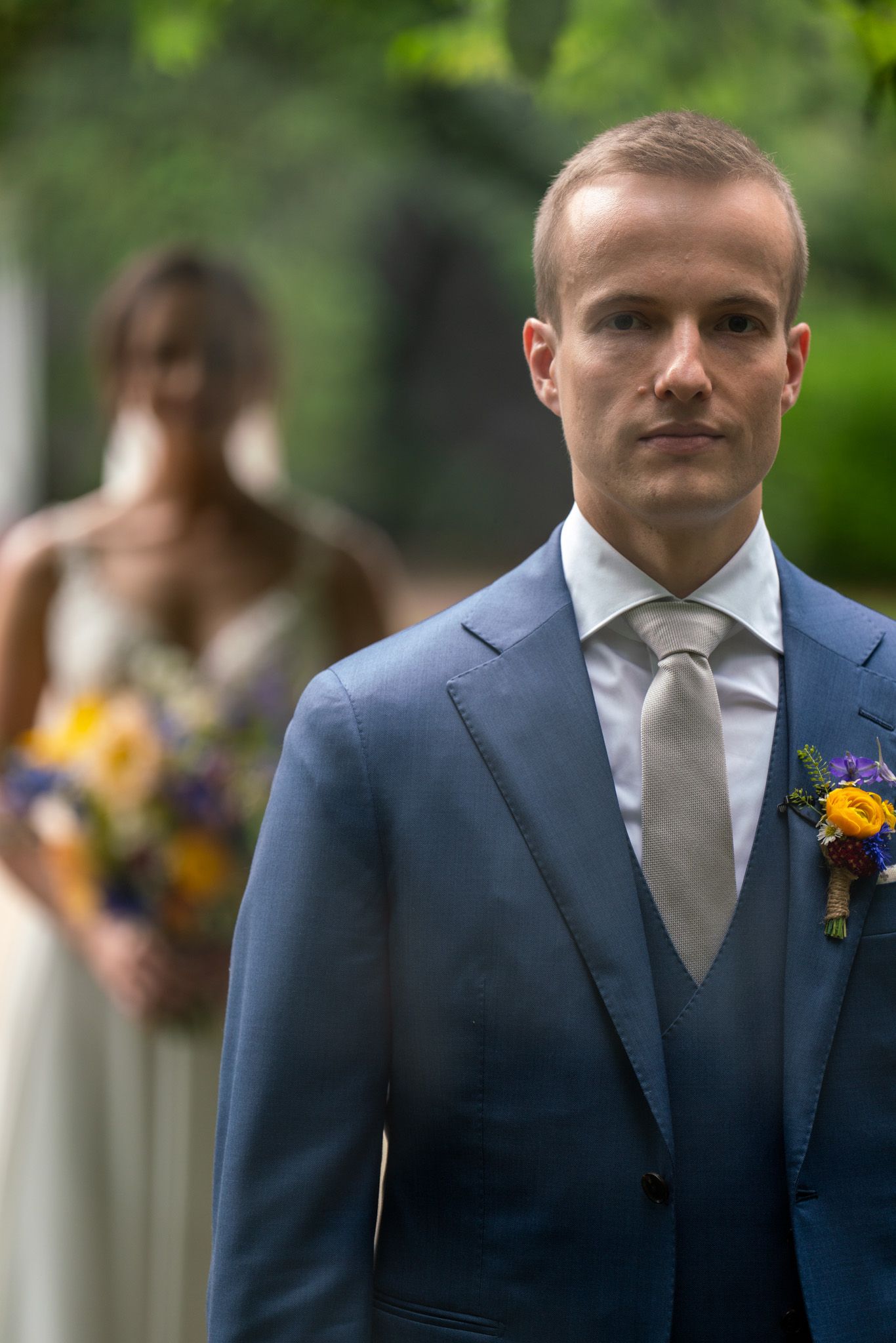 A groom in blue suit with yellow boutonniere during first look with bride visible in background.