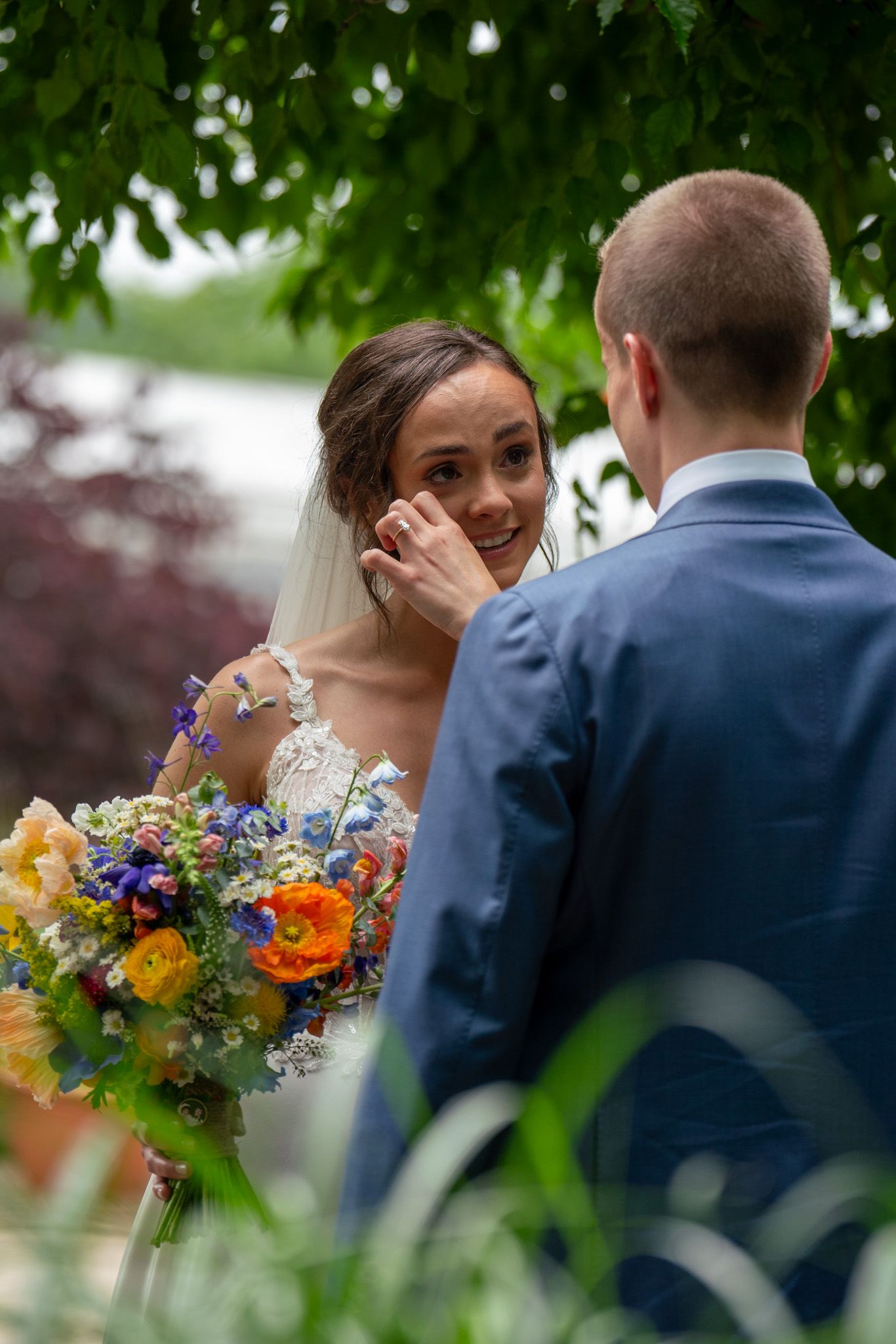 A bride with vibrant wildflower bouquet shares emotional moment with groom under tree canopy.