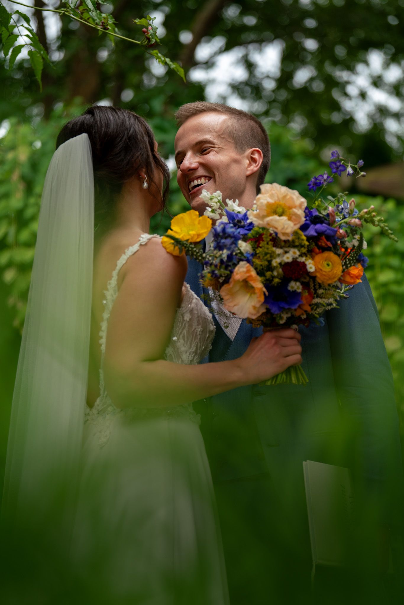 Intimate wedding moment captured through greenery as couple shares smile with colorful bouquet.