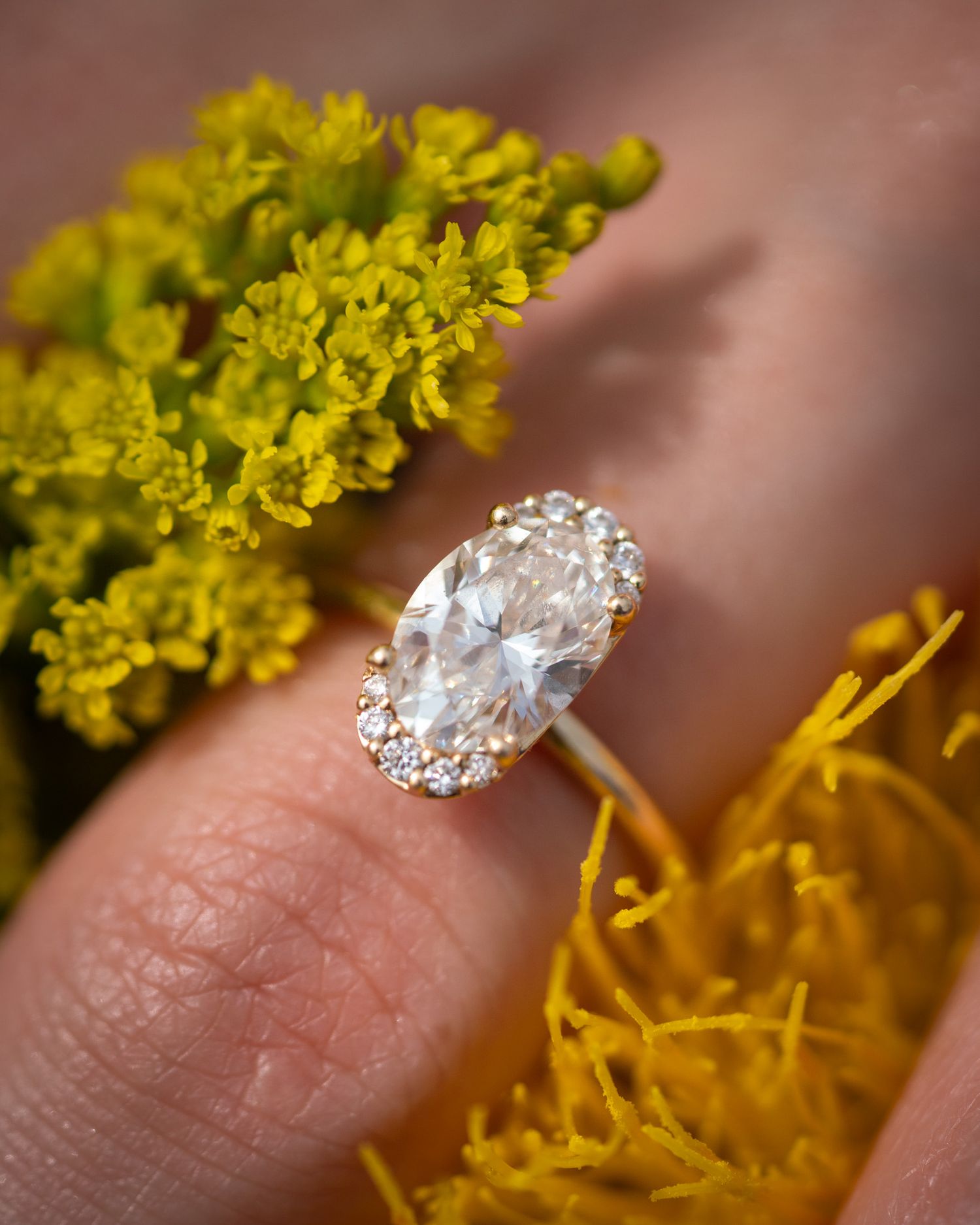 Close-up of oval diamond engagement ring with halo setting against yellow flowers.
