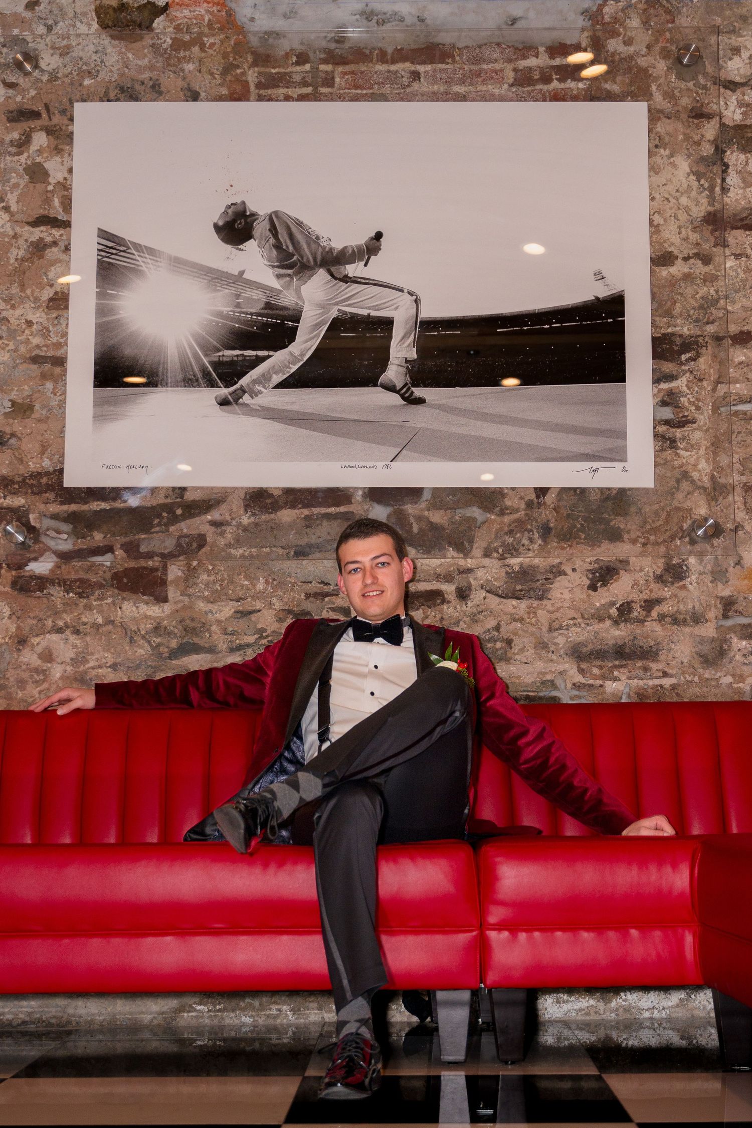 Person in tuxedo lounges on red leather couch beneath iconic Queen photograph.