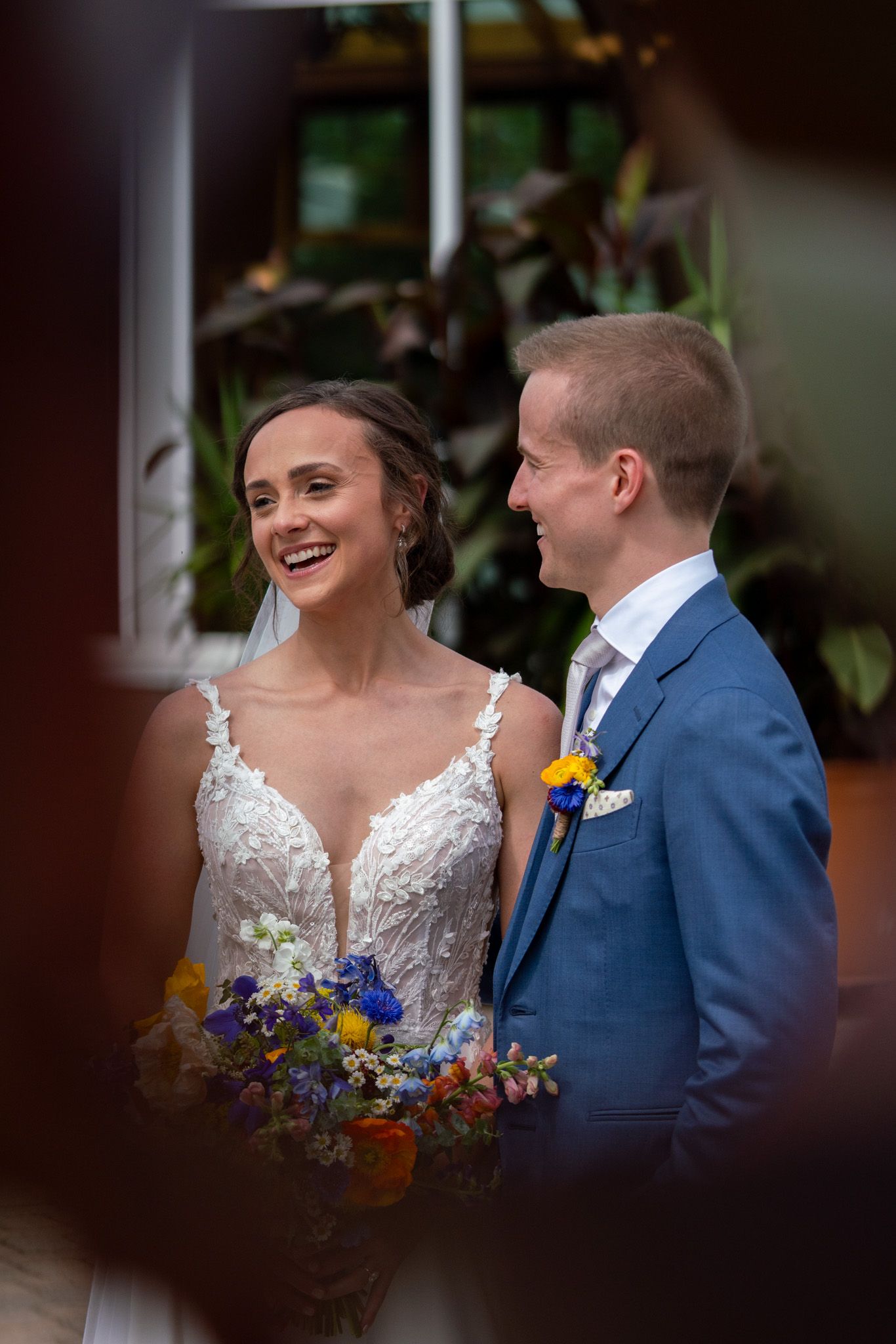 Candid wedding portrait of couple with colorful bouquet framed through window or doorway.