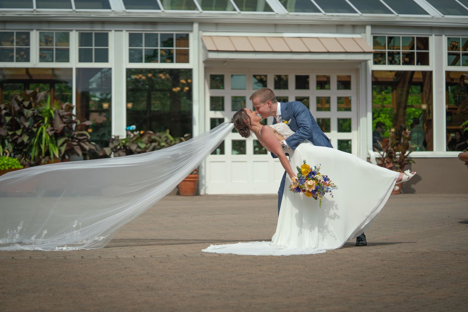 Romantic dip and kiss moment between newlyweds on a patio with a greenhouse backdrop.