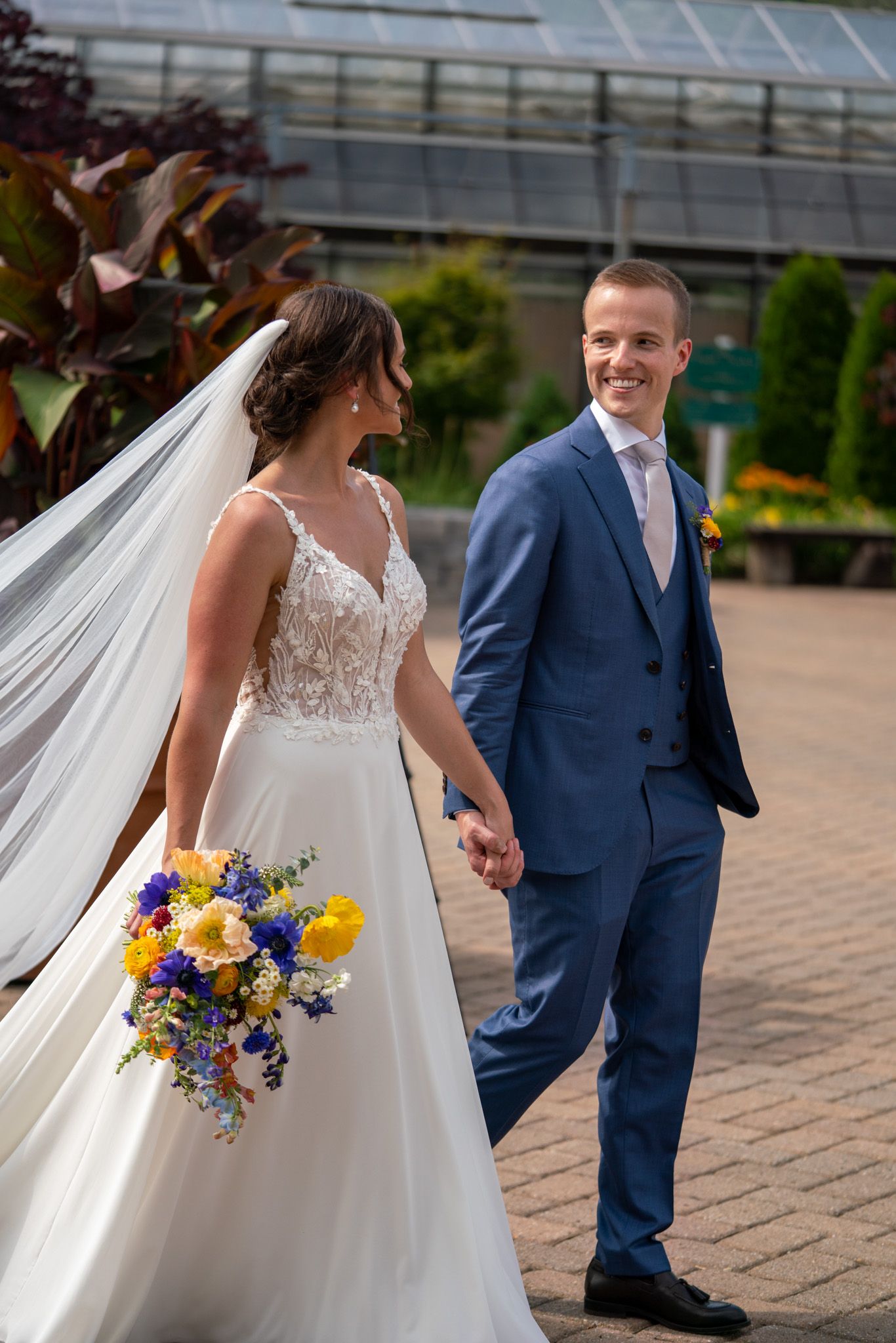 Wedding couple walking outdoors holding hands with colorful summer bouquet near greenhouse.