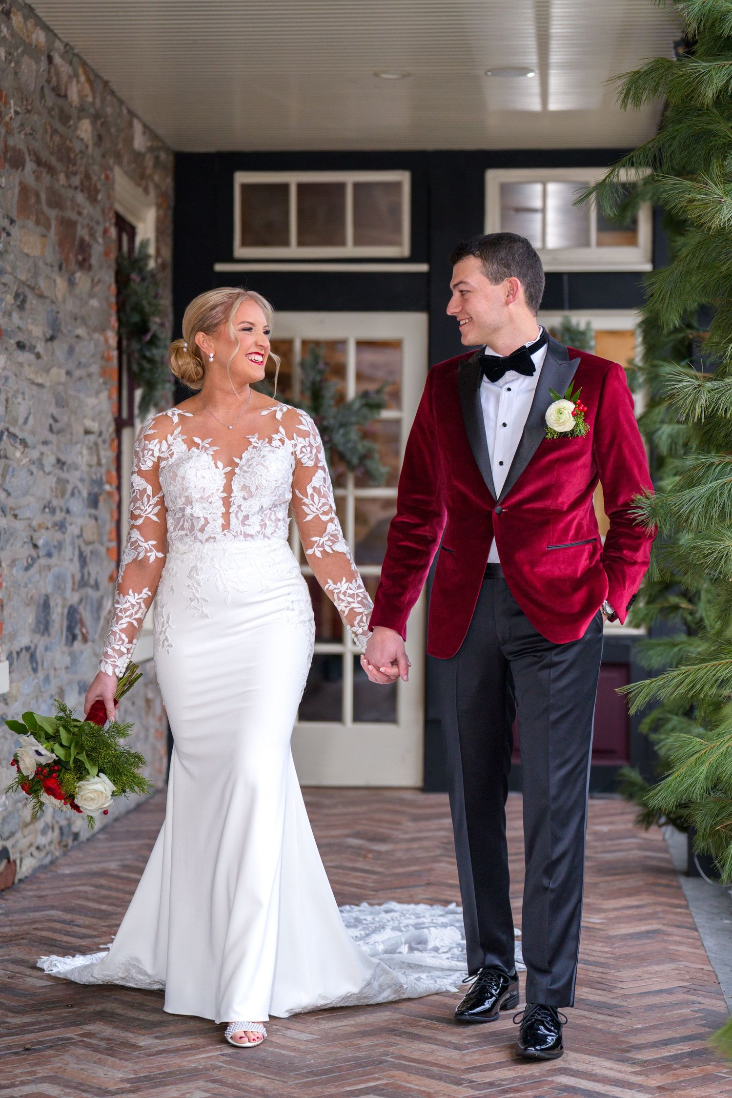 A wedding couple in white lace gown and burgundy velvet tuxedo walk together on a stone porch.