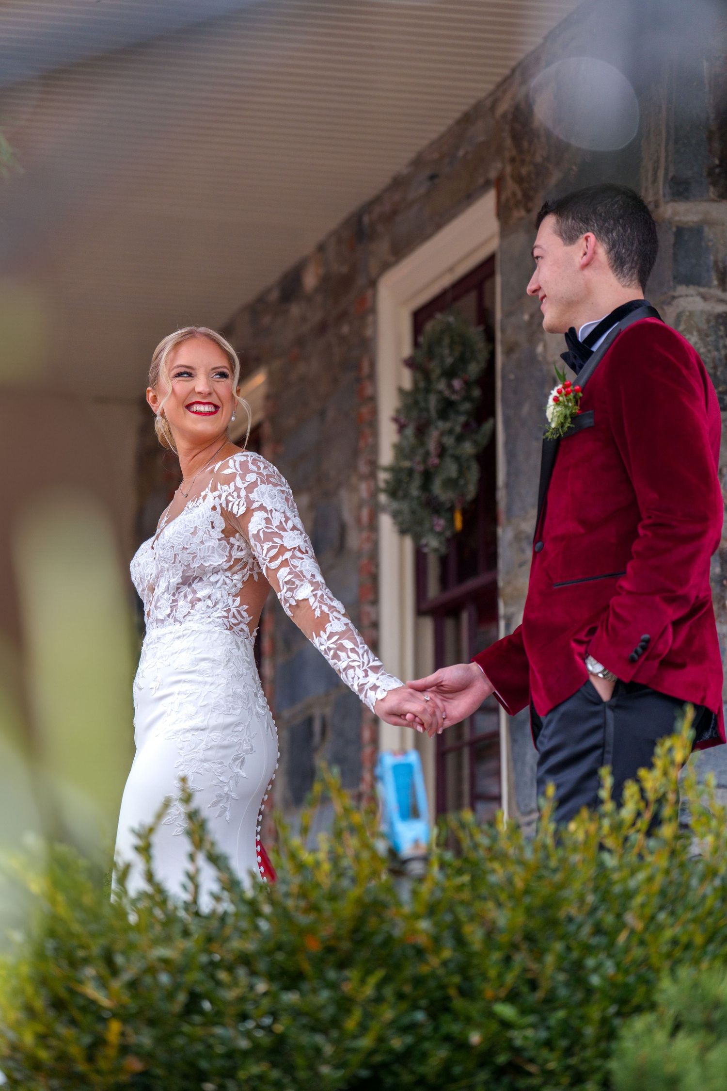 A bride in a lace gown and groom in burgundy velvet stand together at a stone entrance during their winter wedding ceremony.