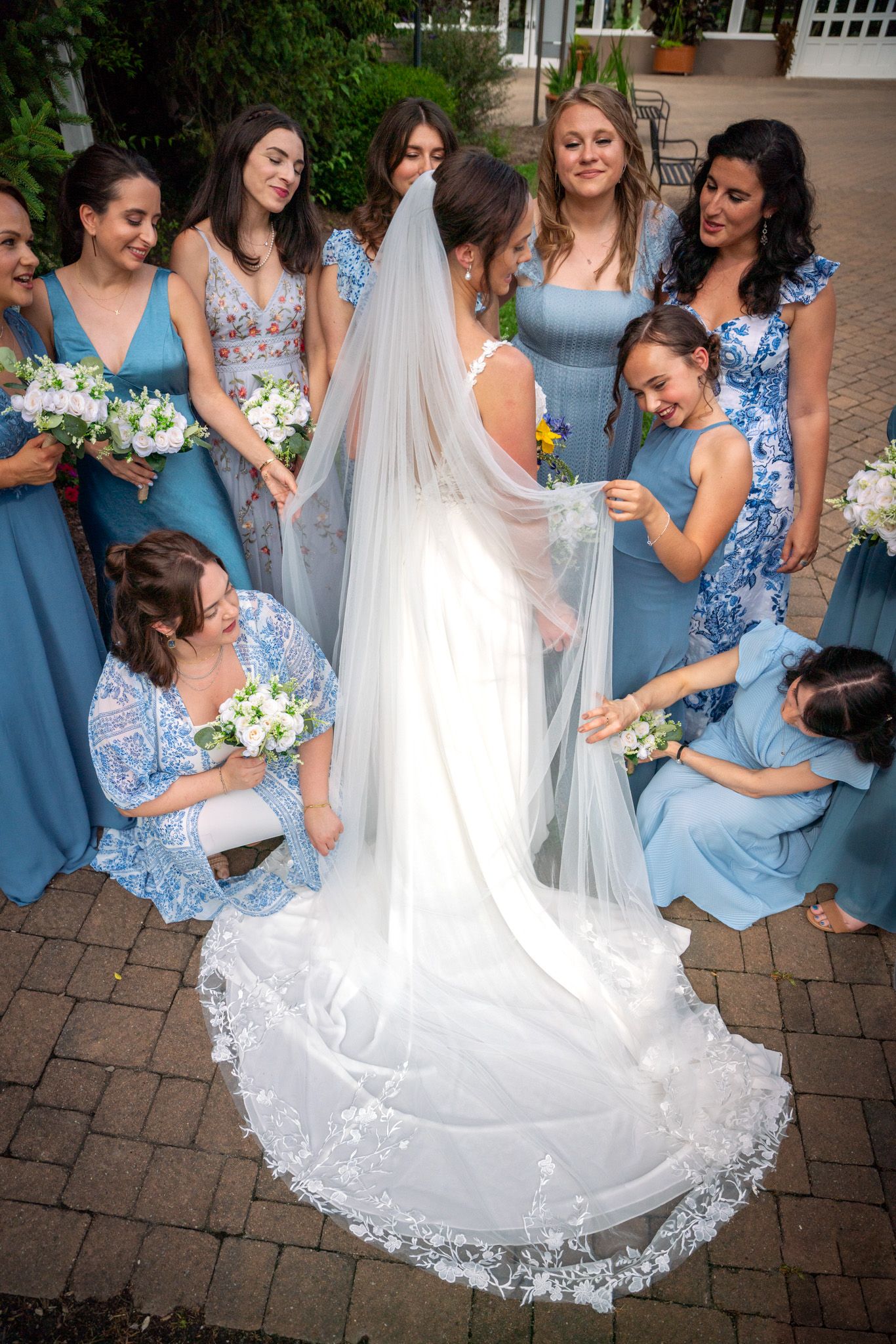 Bridesmaids in blue dresses gather around to admire a long white wedding veil on brick patio.