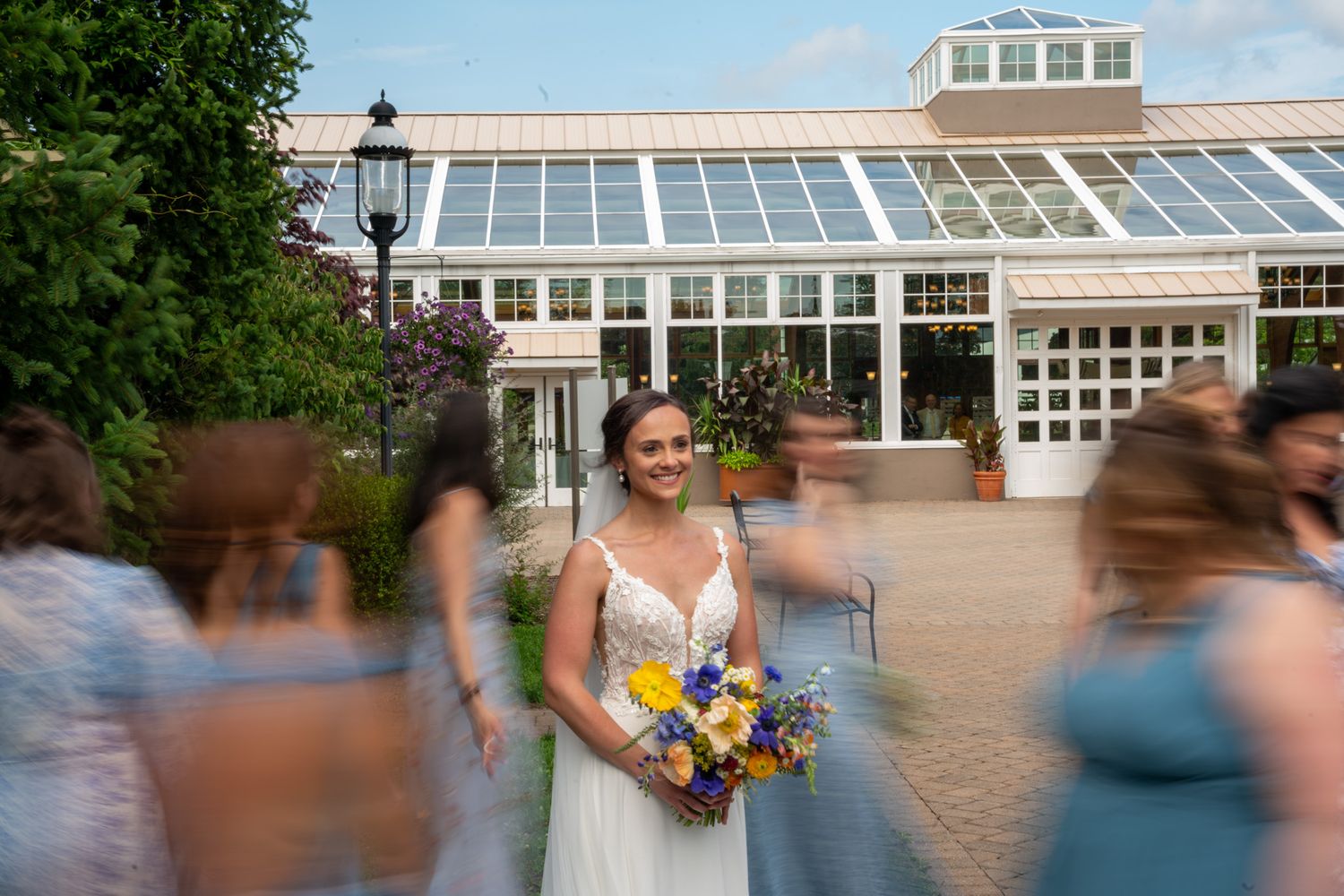 Bride with vibrant yellow and purple bouquet standing outside glass conservatory.