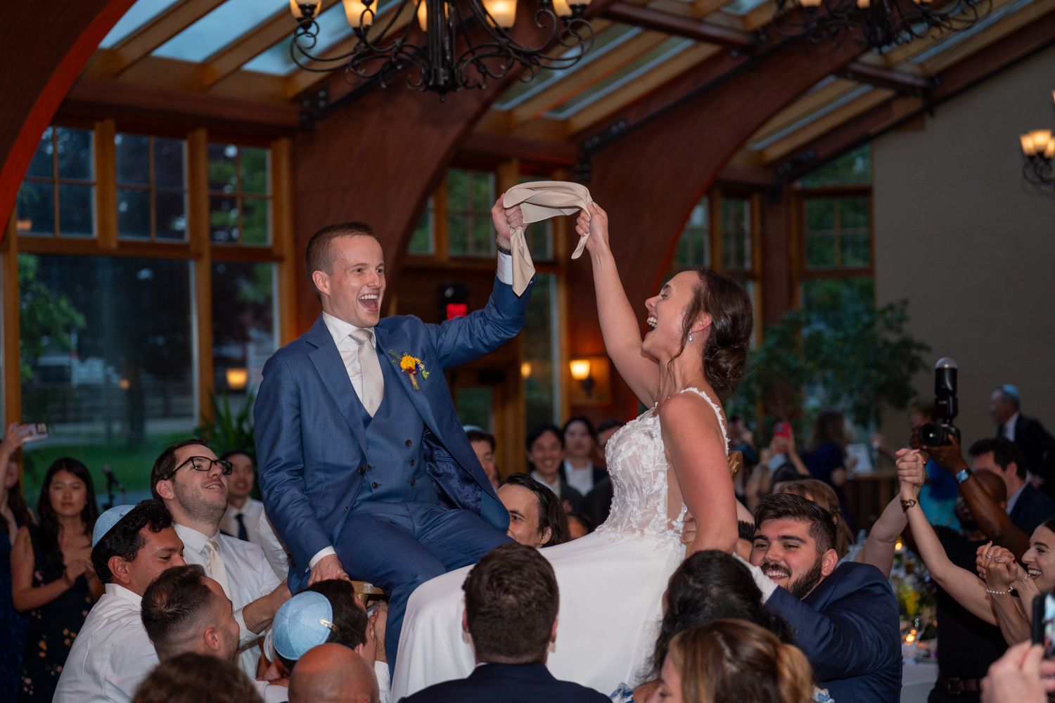 Newlyweds being lifted on chairs during traditional wedding dance celebration.