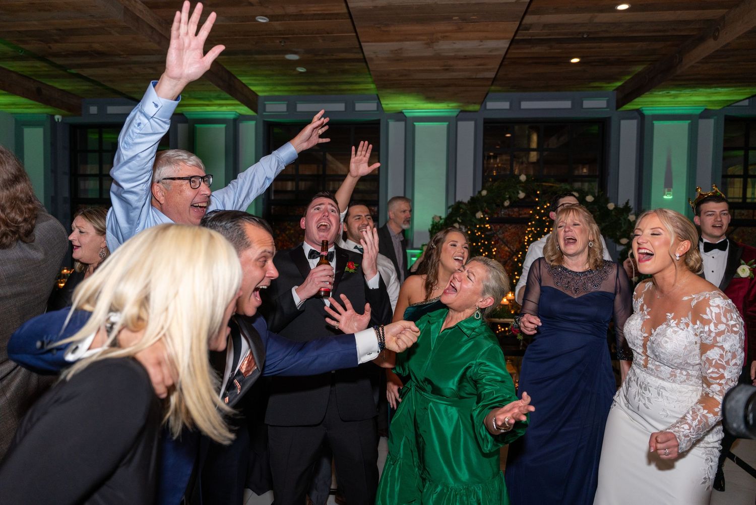 Wedding reception guests dancing and celebrating with raised hands under green lighting.