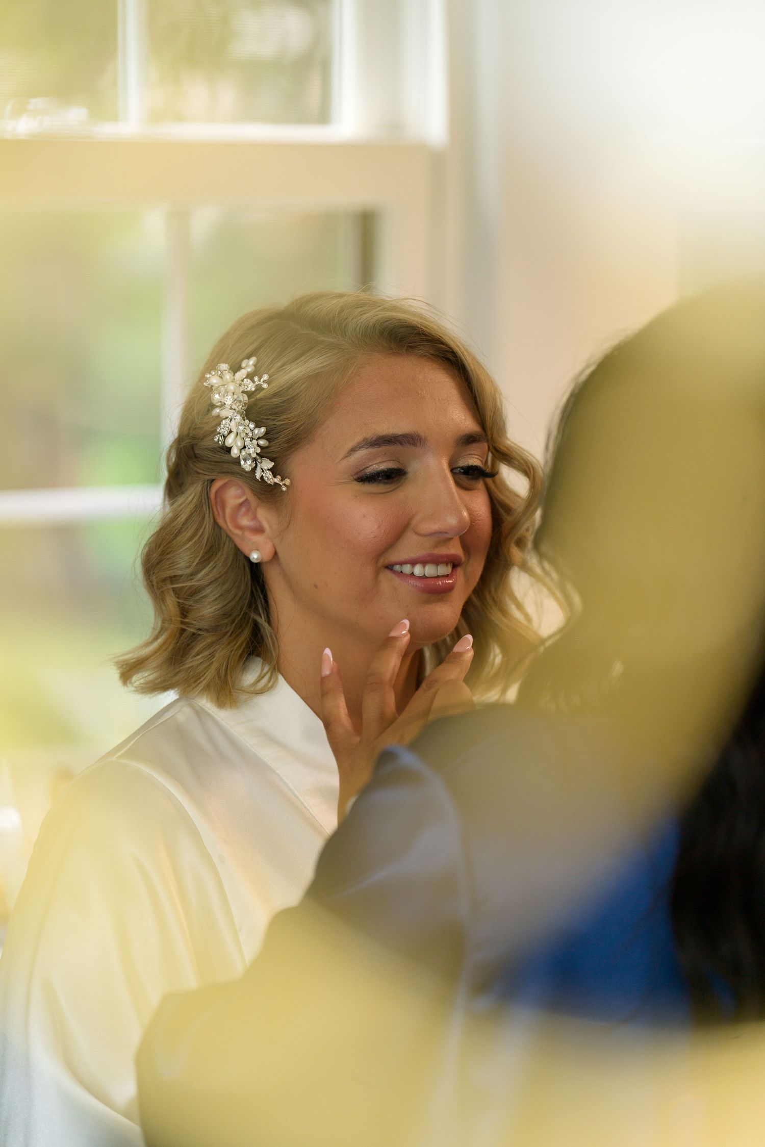 Elegant bride smiles while getting ready in a white robe, wearing a decorative crystal hair clip.