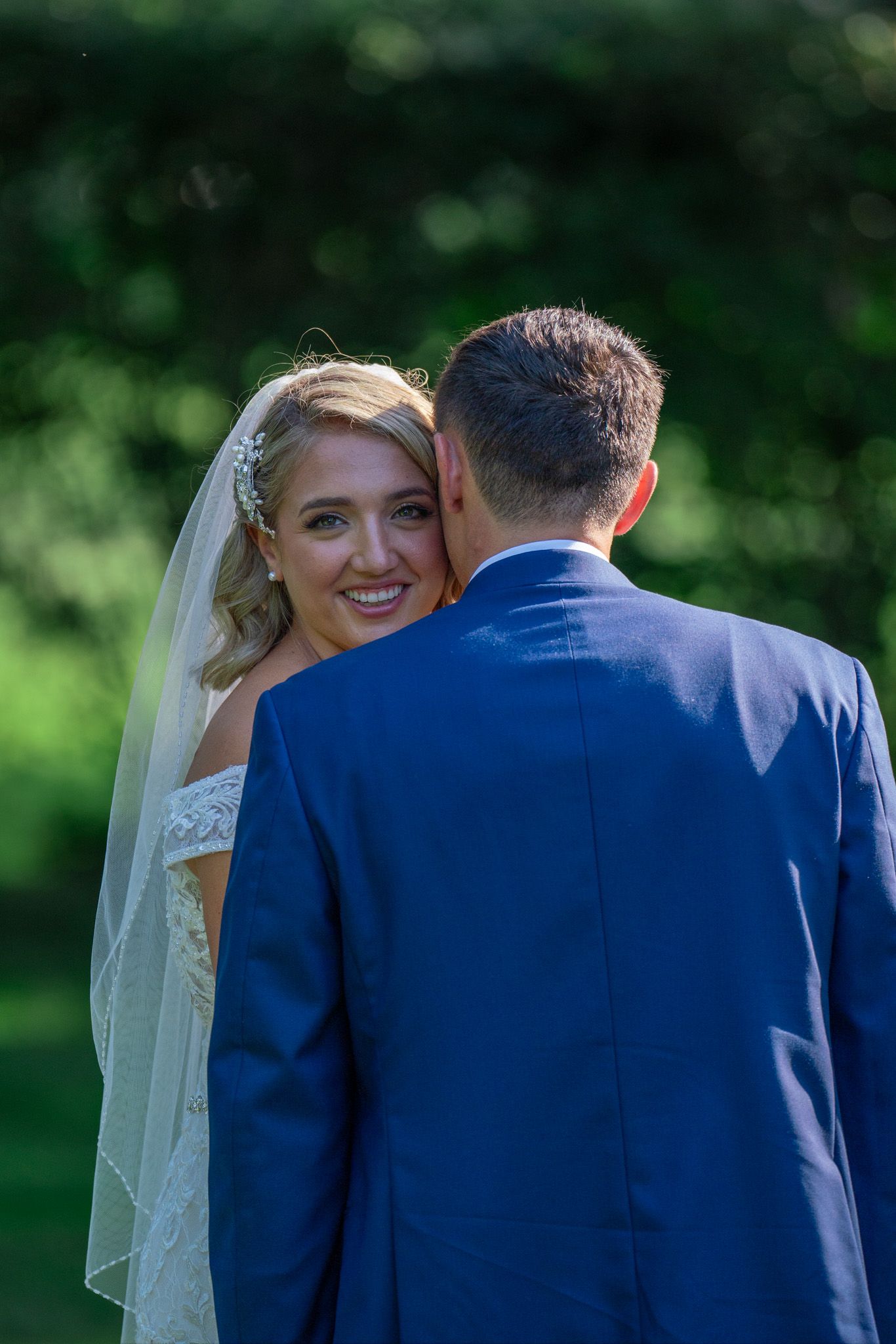 Bride smiles radiantly while standing with groom in navy suit against green foliage.