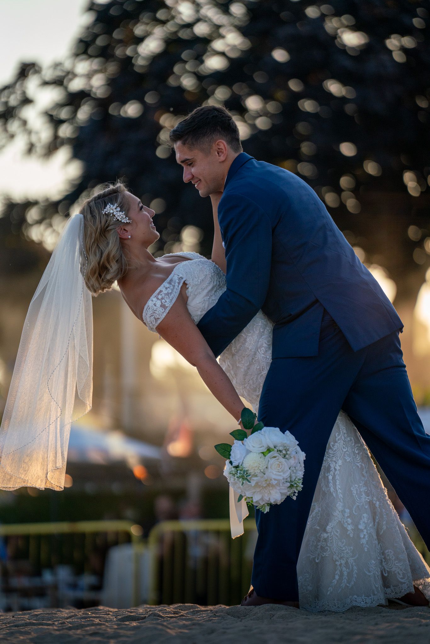 Artistic wedding photo shows romantic dip pose against bokeh tree backdrop at sunset.