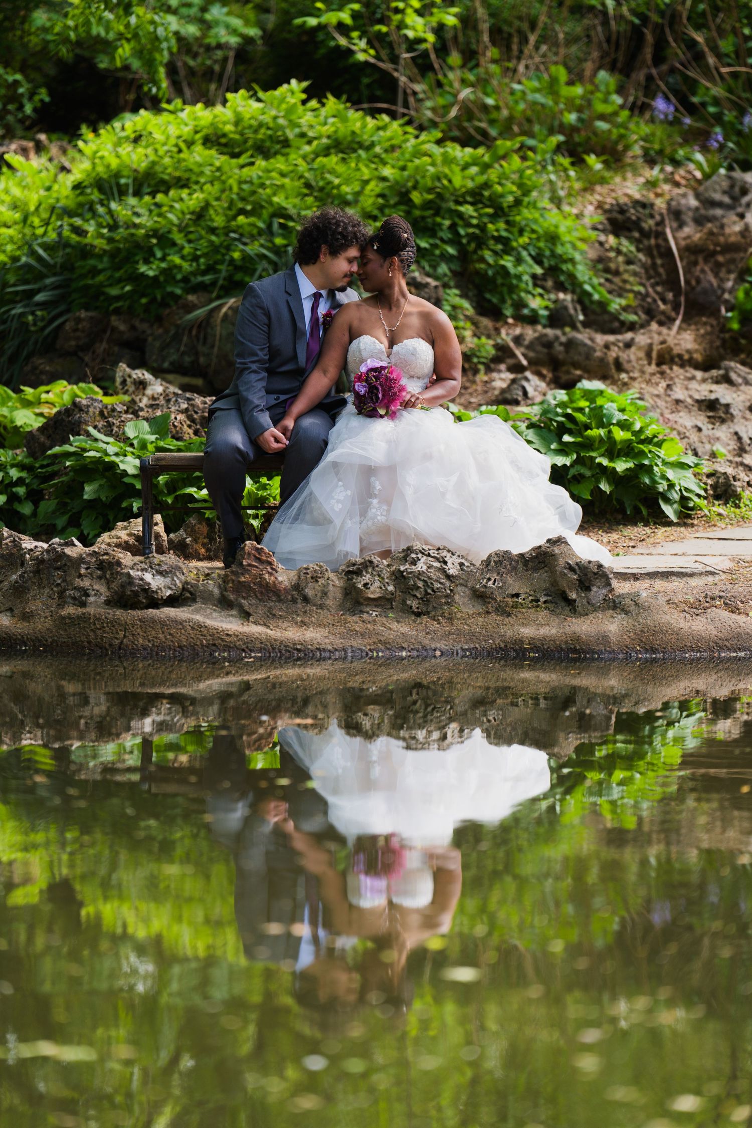 A romantic wedding reflection photo by a pond with lush greenery in the background.
