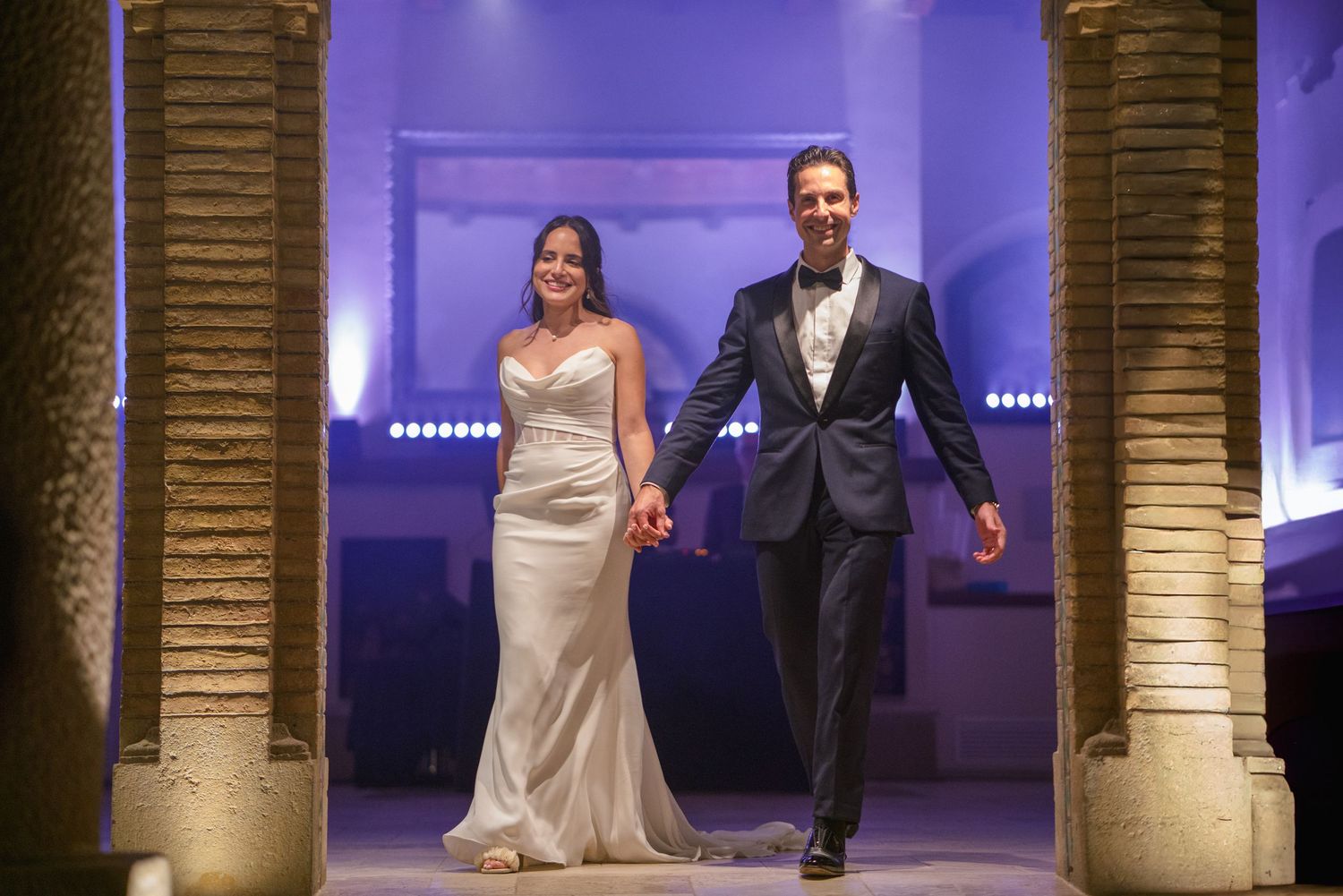 A couple makes their grand entrance through stone columns with purple lighting at their wedding reception.