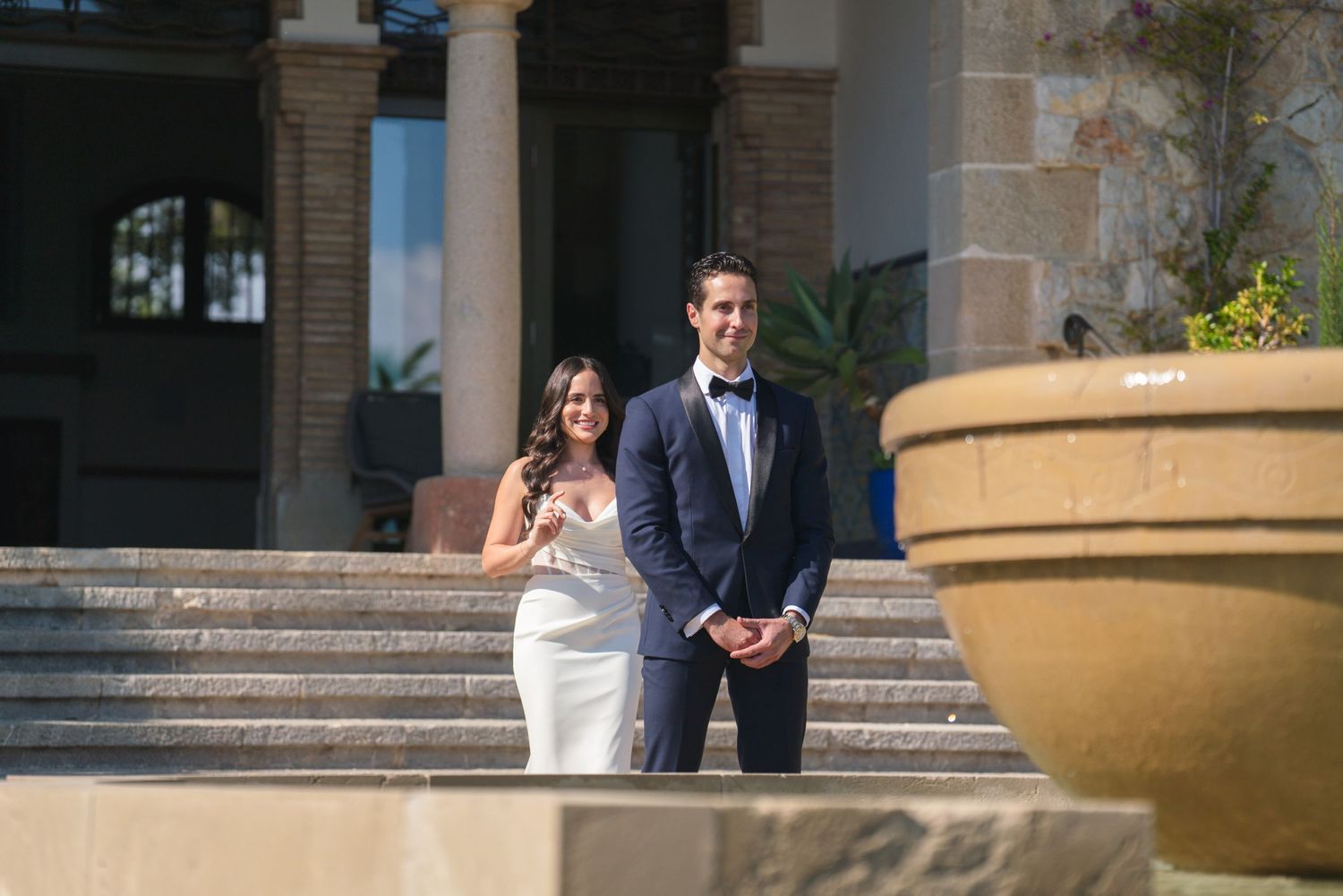 A wedding couple stands on elegant stone steps in formal attire.