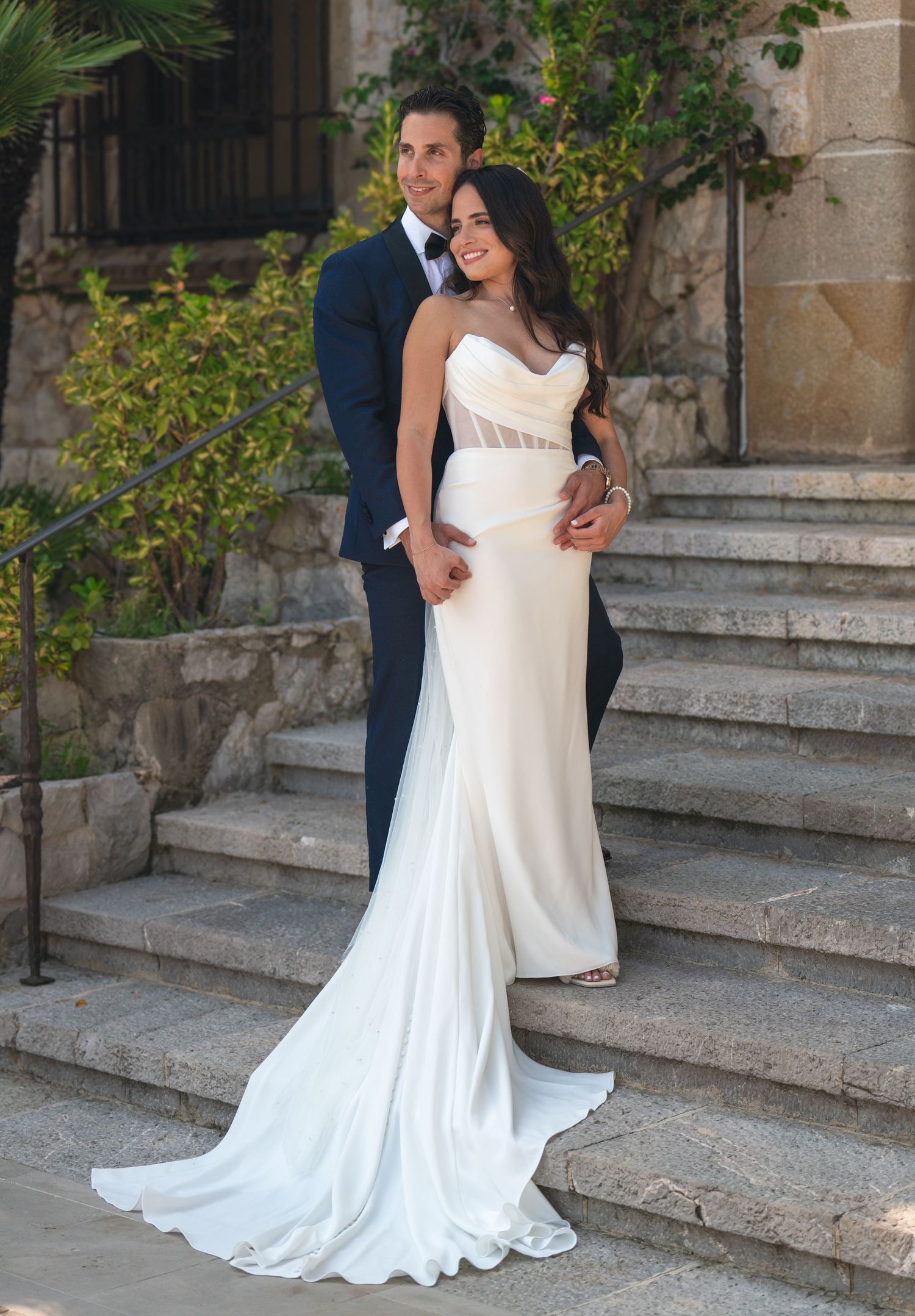 A wedding couple poses on stone steps with bride wearing an elegant fitted white gown.