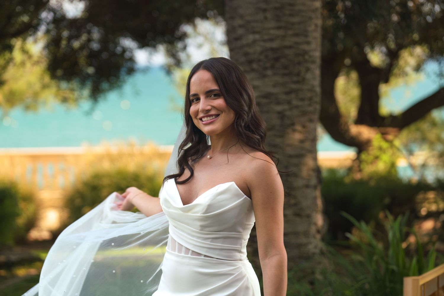 A bride in a stunning white strapless gown smiles while holding her veil outdoors.