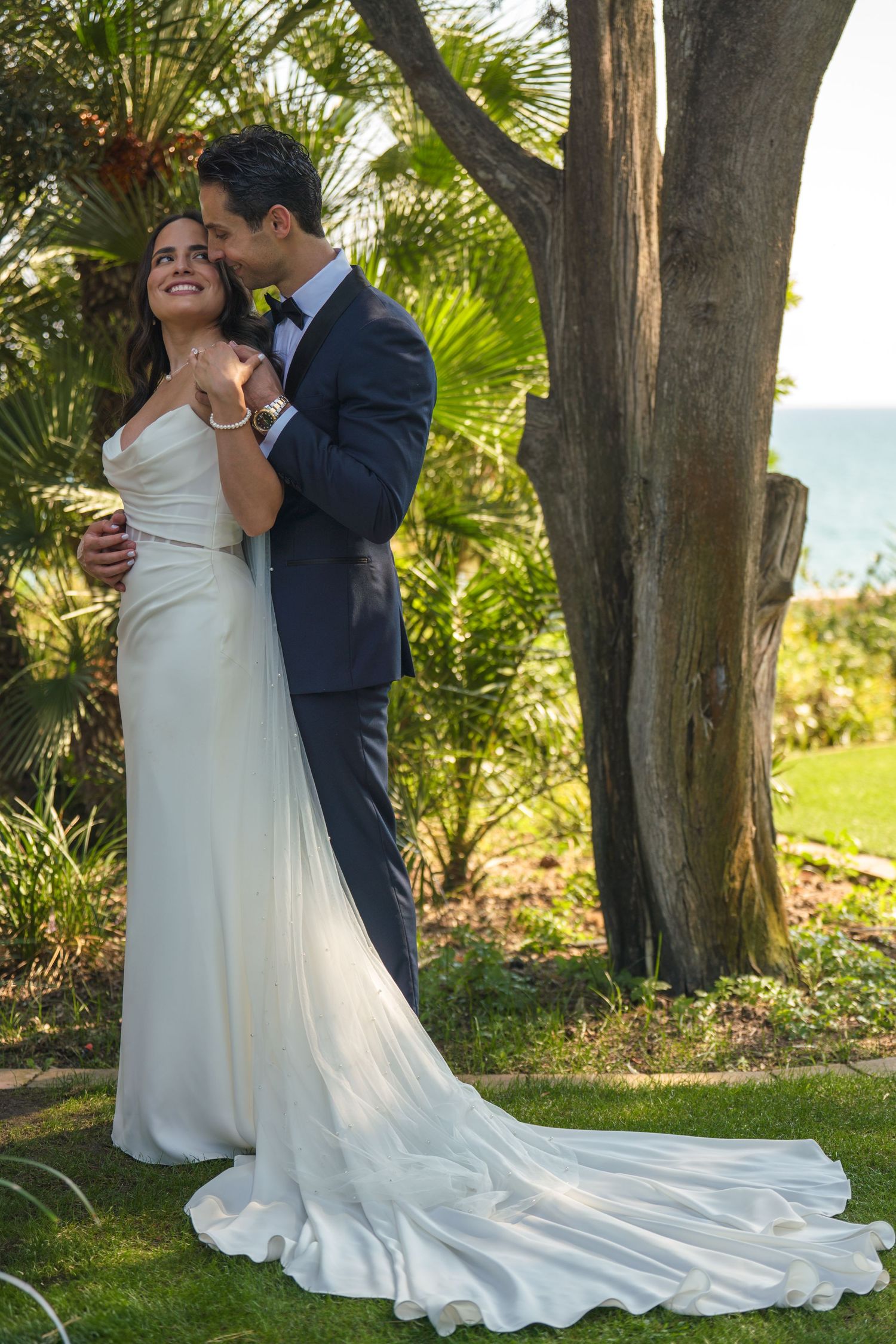 A romantic wedding photo captured in a lush tropical garden setting with ocean views.