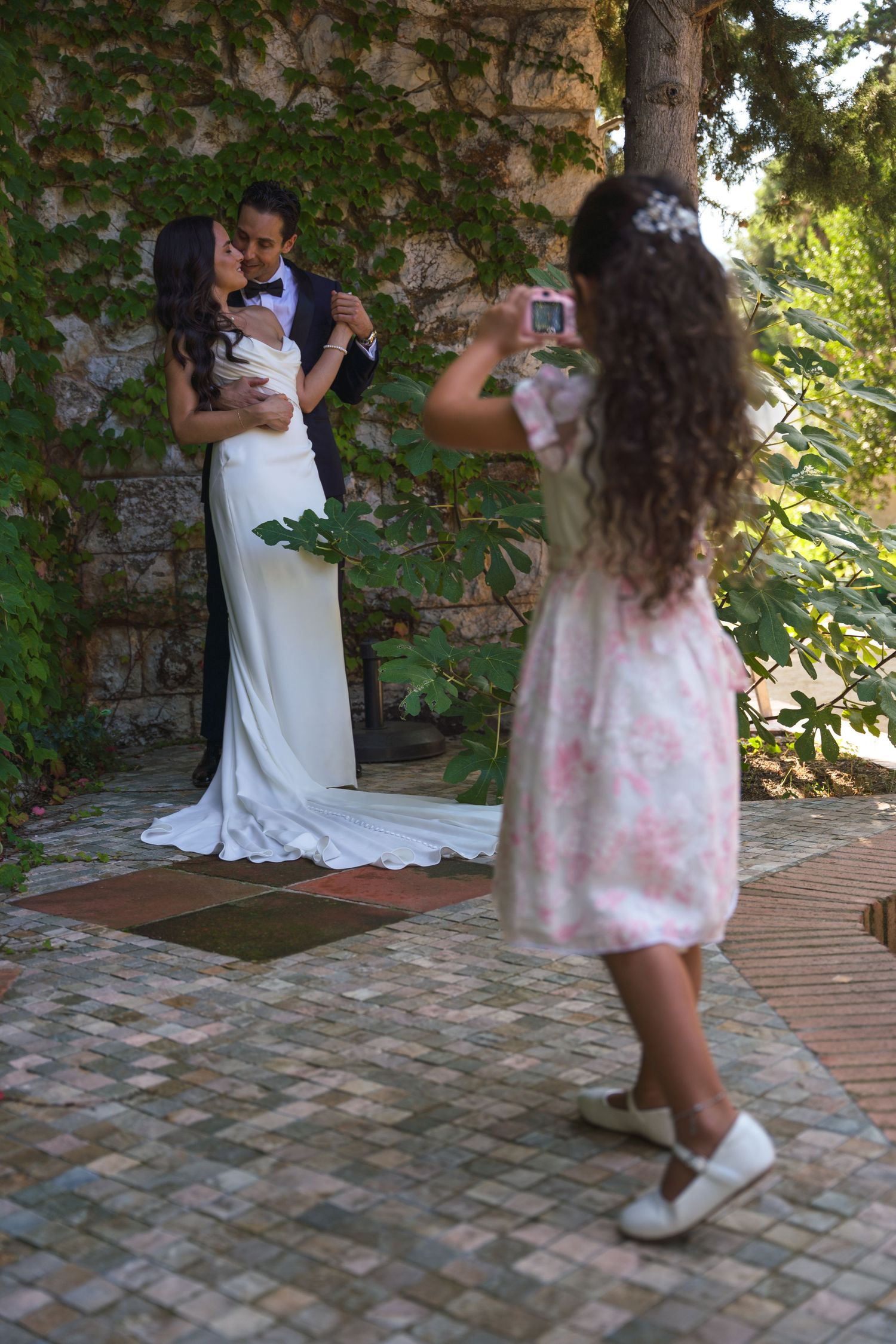 A photographer captures newlyweds posing against a vine-covered wall.