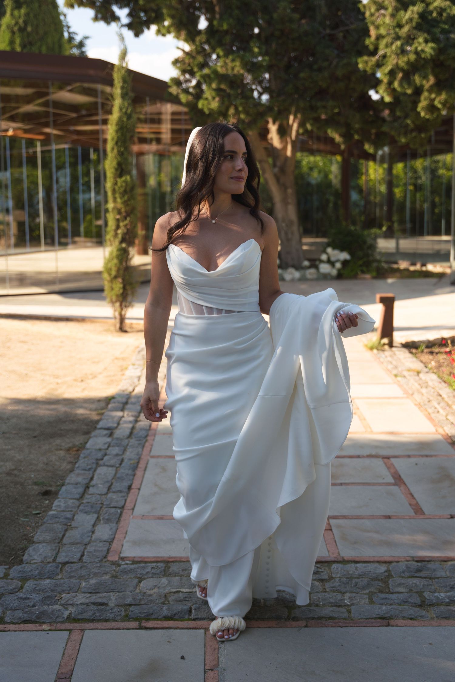 A bride in an elegant white fitted dress walks gracefully along a stone pathway.