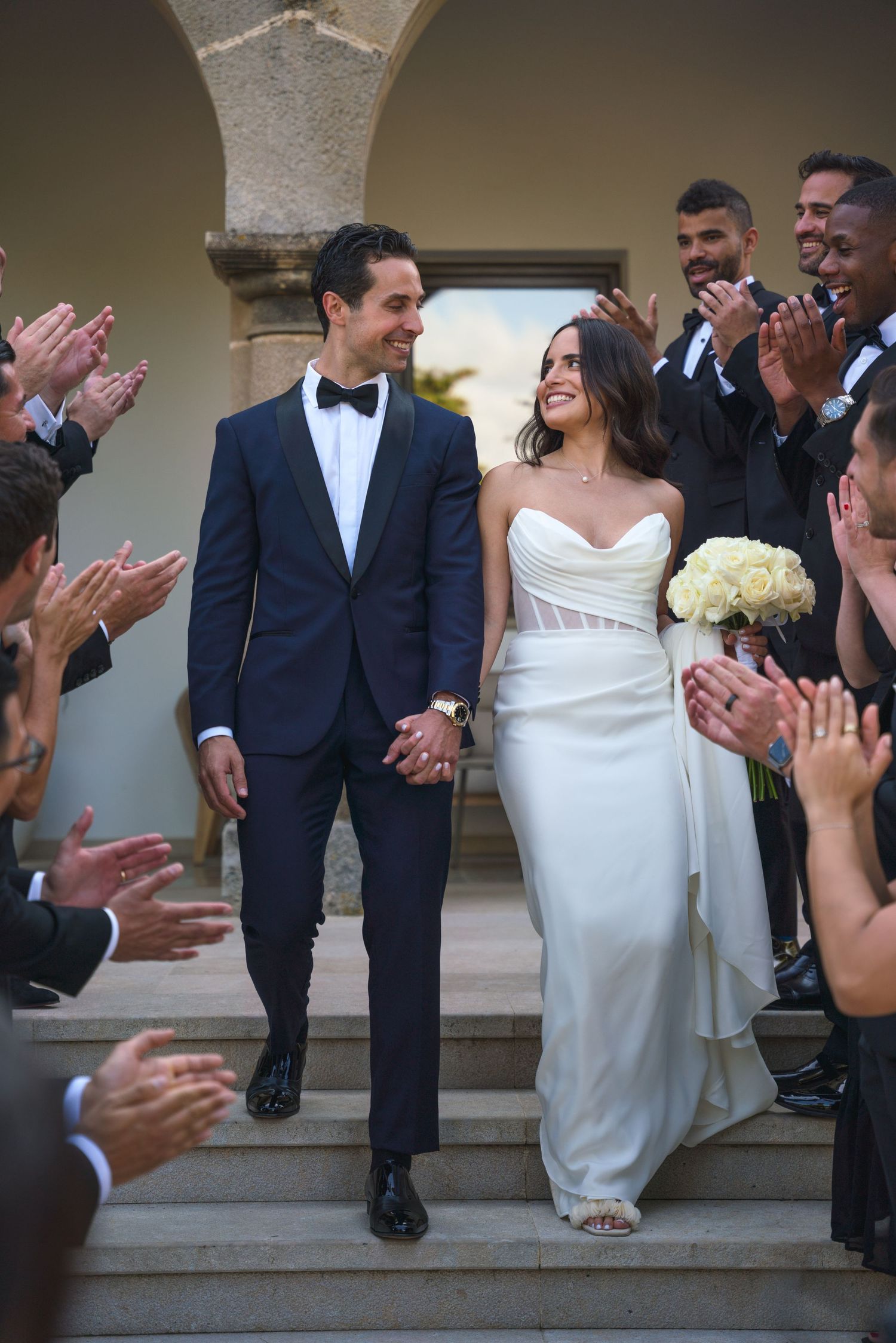 Newlyweds descending stone steps as wedding guests applaud along both sides in an elegant outdoor setting.