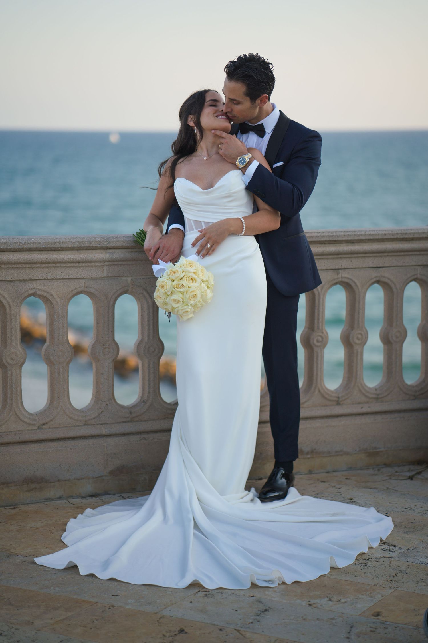 Romantic wedding couple embracing on a seaside balcony with white bouquet and flowing bridal gown.