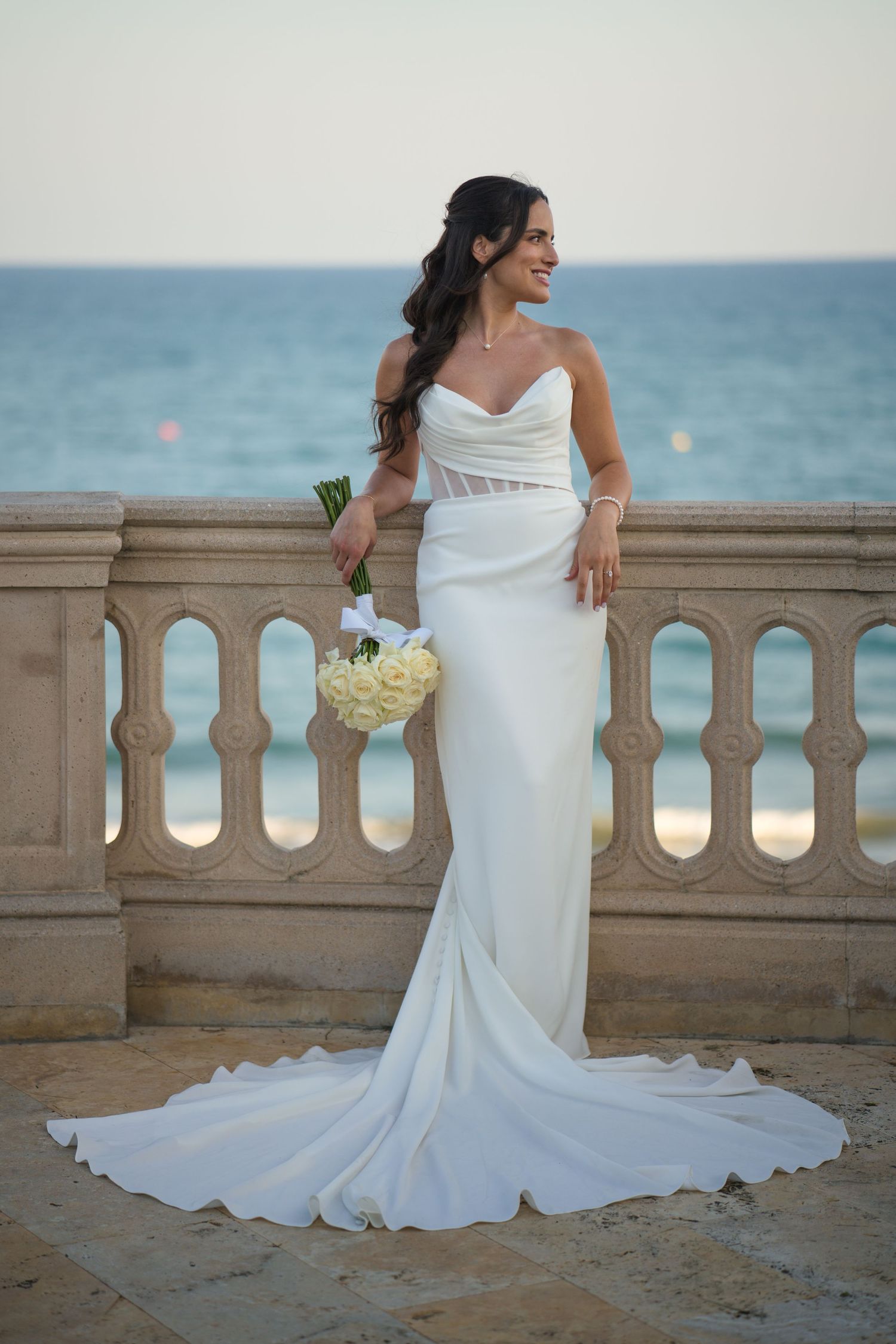 A bride in an elegant white mermaid dress poses on a seaside balcony with a bouquet.
