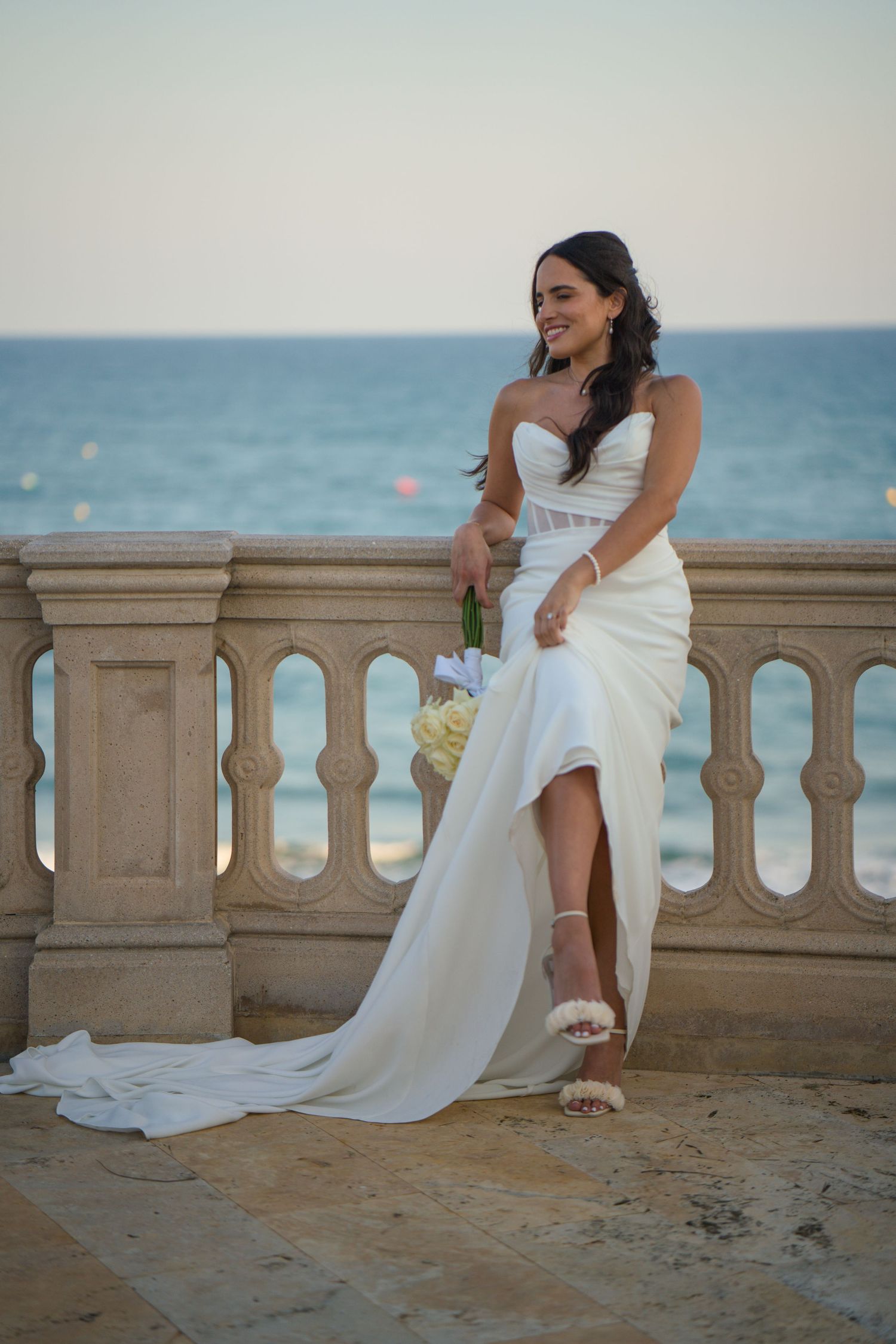A bride in a flowing white dress poses elegantly on a seaside terrace.