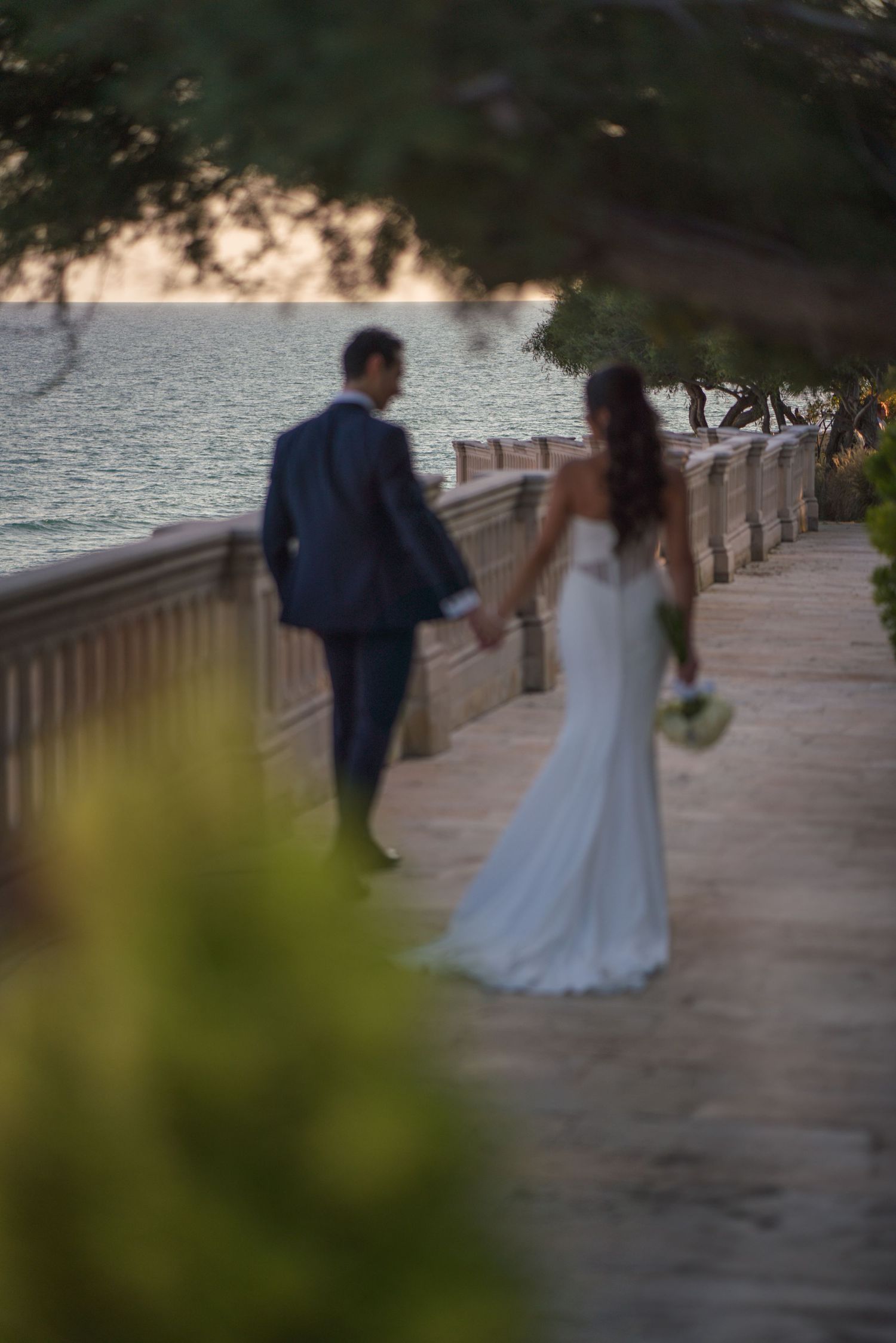 Couple walking along oceanfront path with bride in flowing white dress captured through foliage.