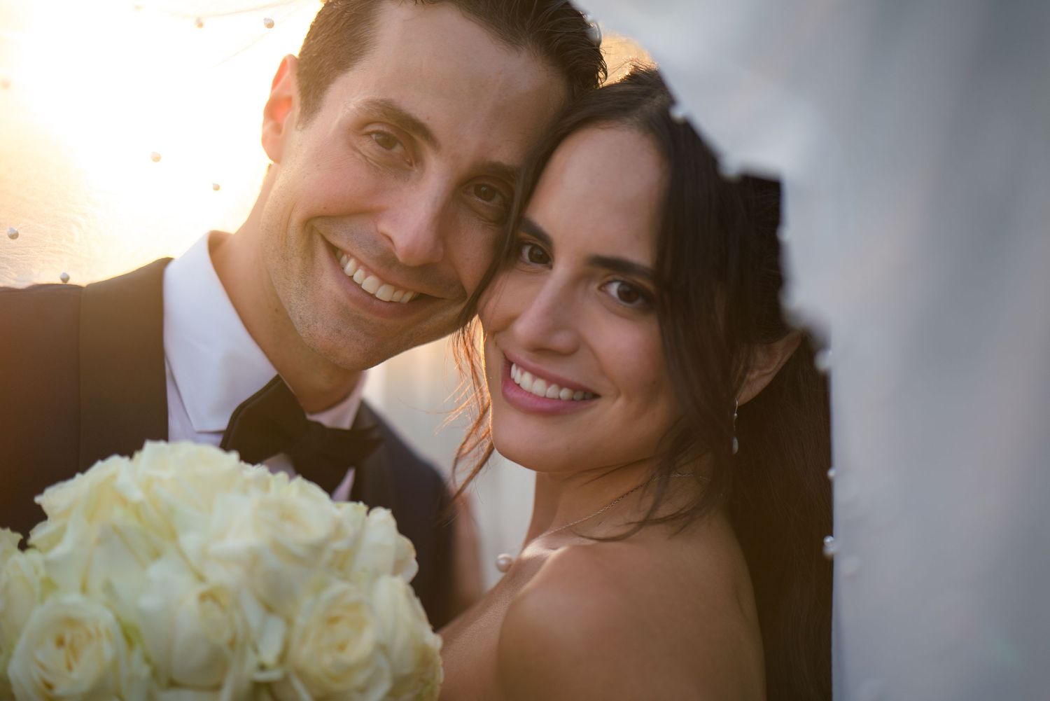 A romantic wedding portrait of a couple holding a white rose bouquet with soft sunset lighting.