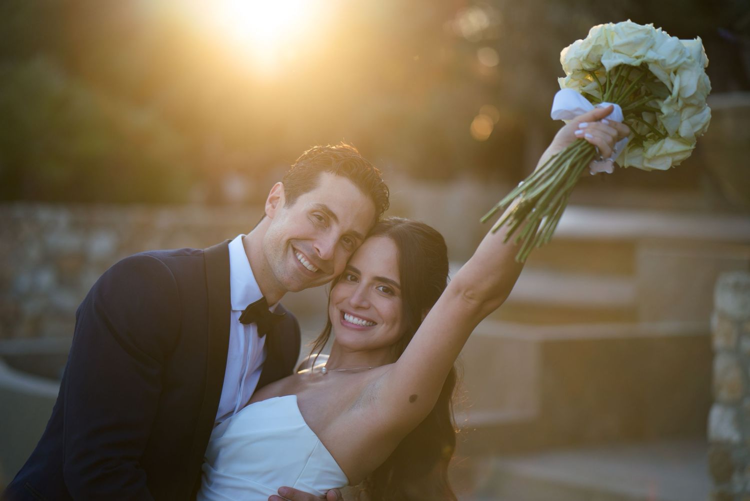 A wedding couple poses during golden hour sunset with raised bouquet.