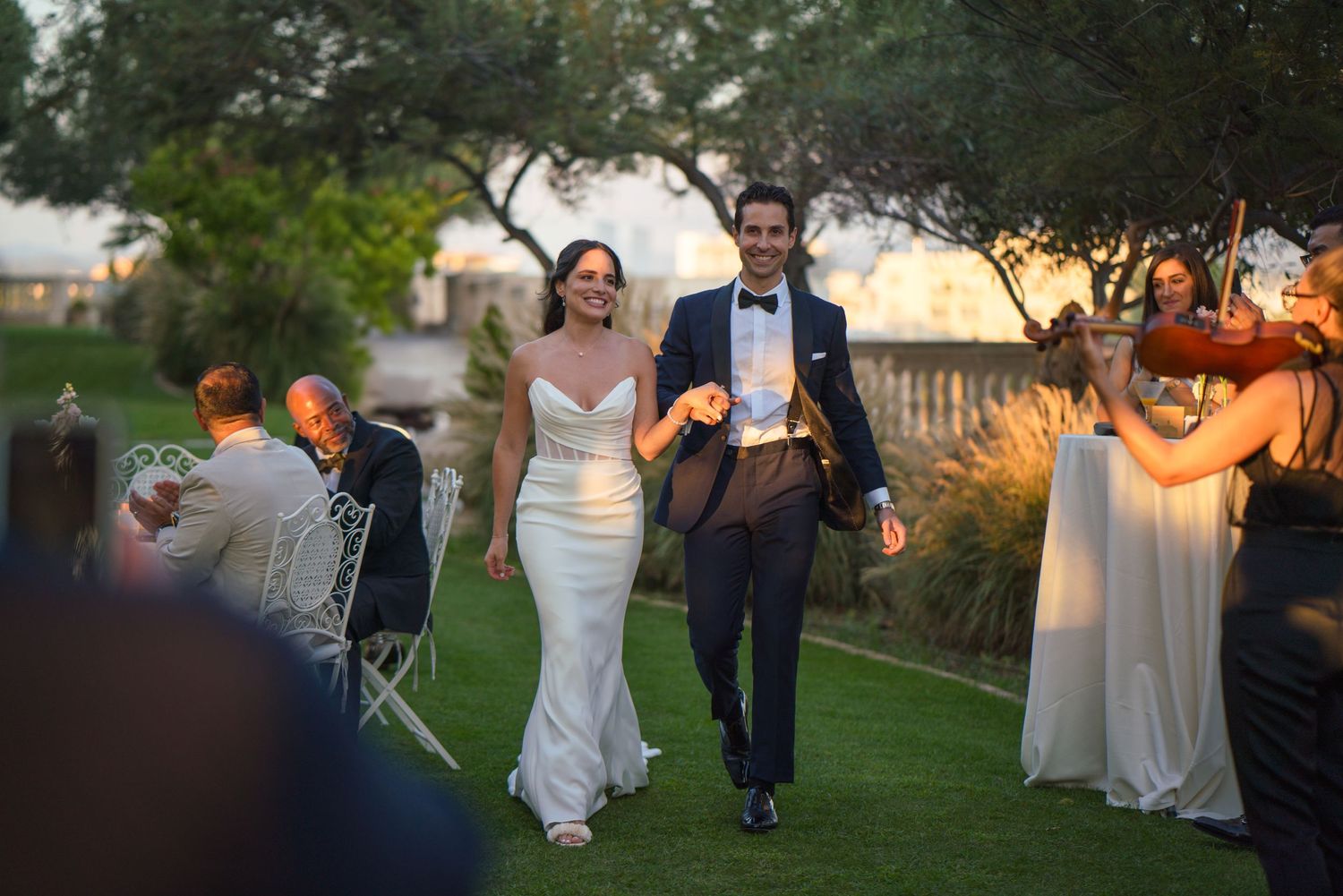 Wedding couple walking through garden reception as guests celebrate in background.