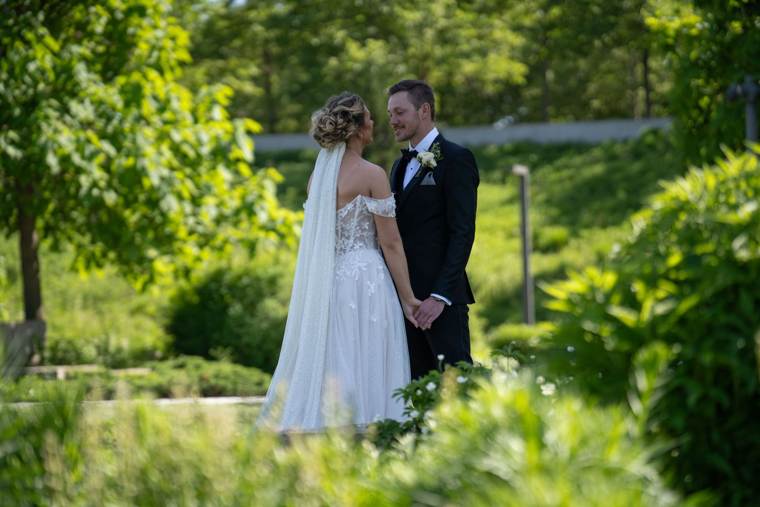 A couple in wedding attire stands together in a lush garden setting with greenery in the background.