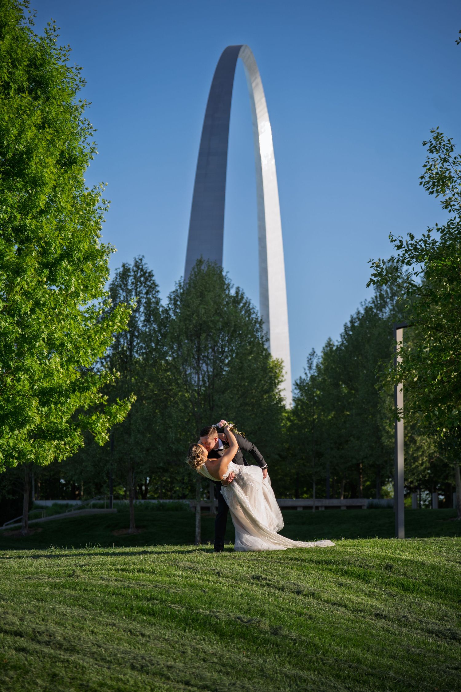 A wedding couple shares a romantic moment on a grassy hill with the iconic St. Louis Gateway Arch in the background.