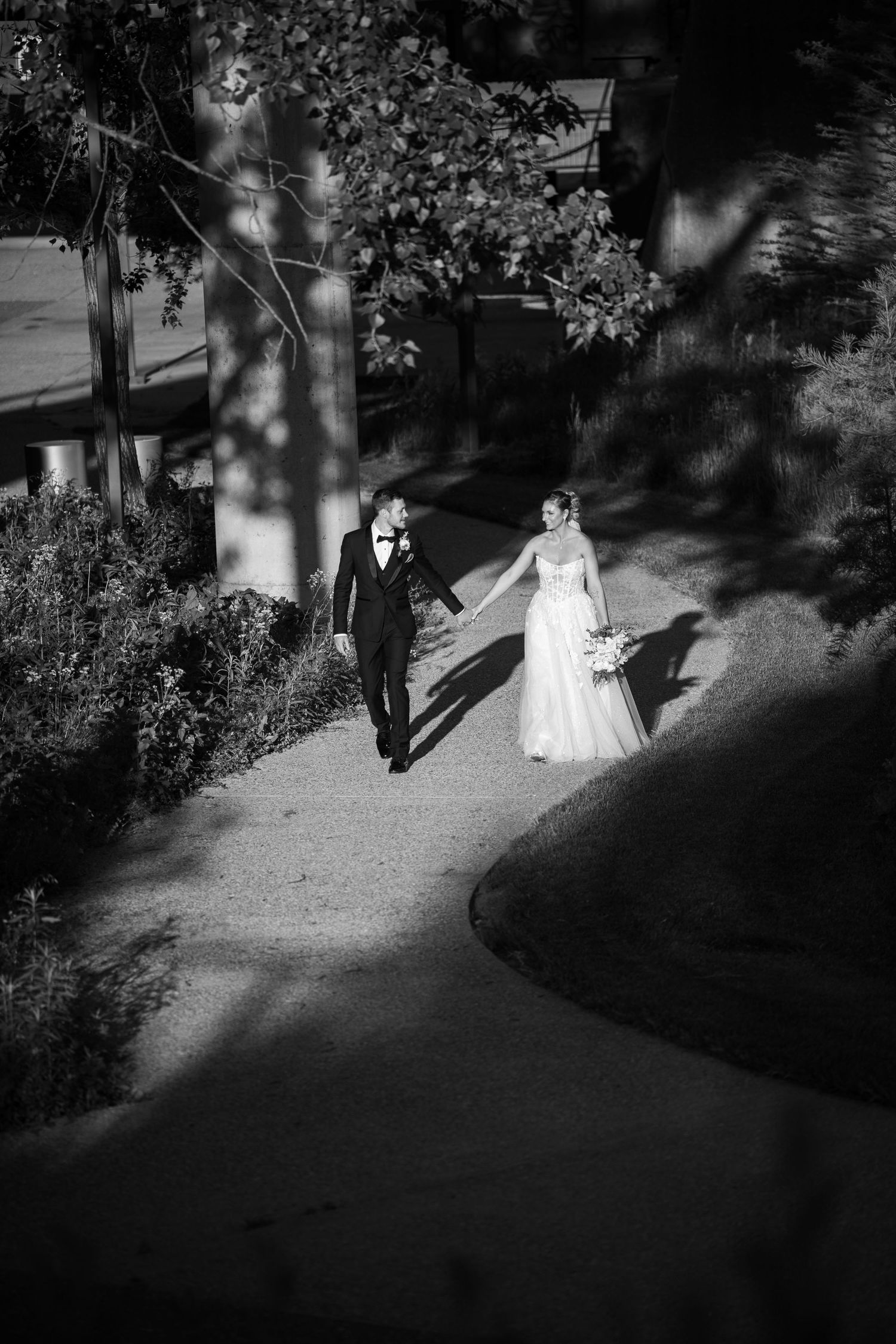 Black and white wedding photos capture a couple walking along a garden path with dappled sunlight and shadows.