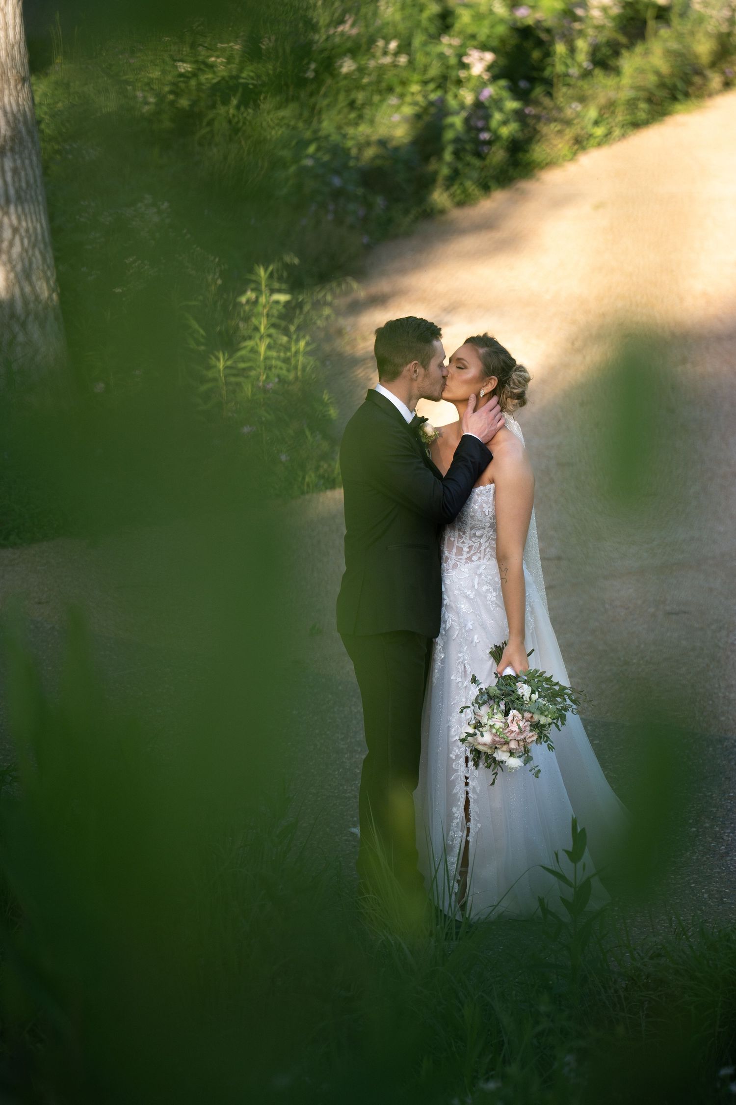 A romantic wedding couple shares an intimate moment on a sunlit garden path framed by green foliage.