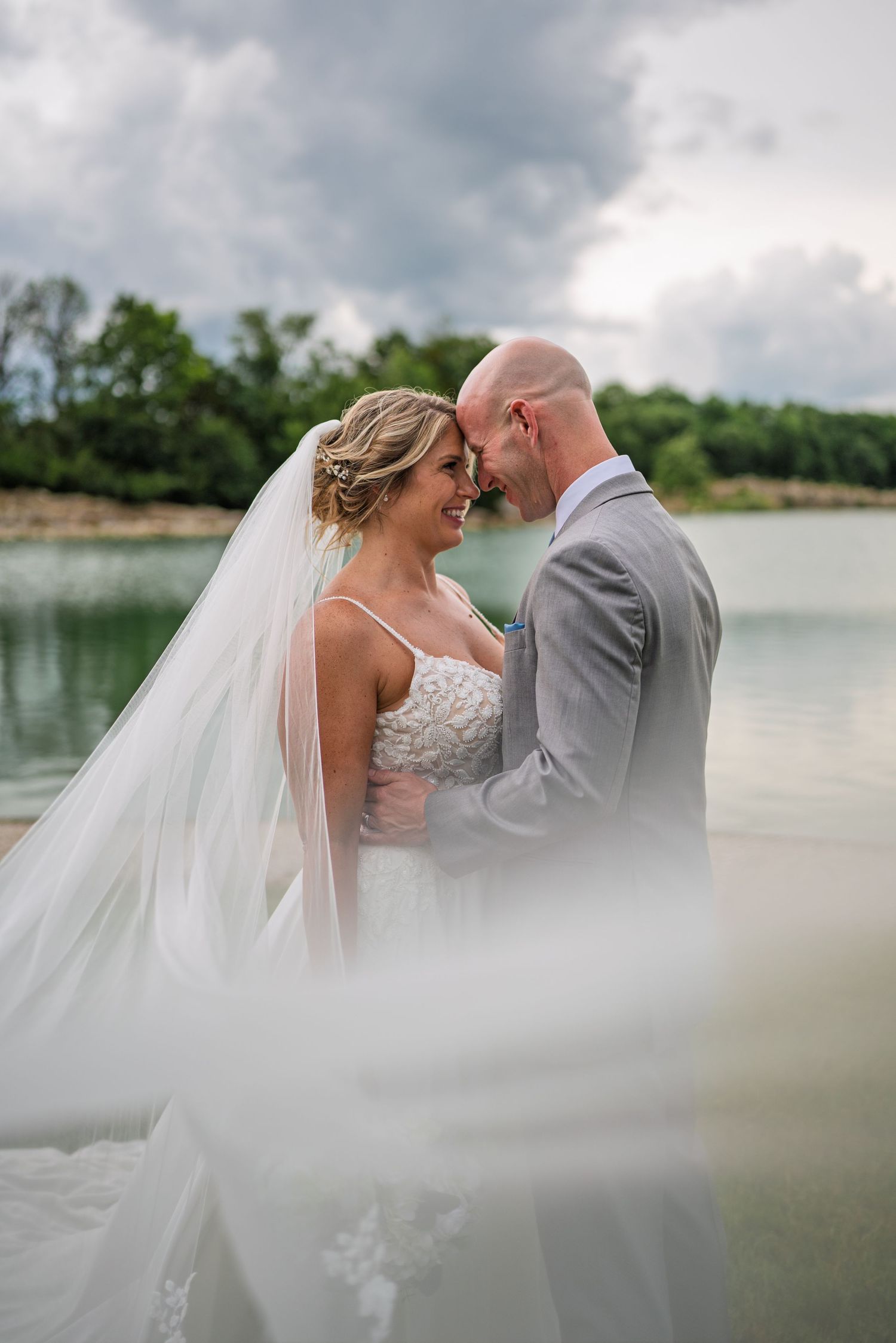 A romantic wedding couple embraces by a tranquil lake with flowing veil in foreground, set against cloudy skies.