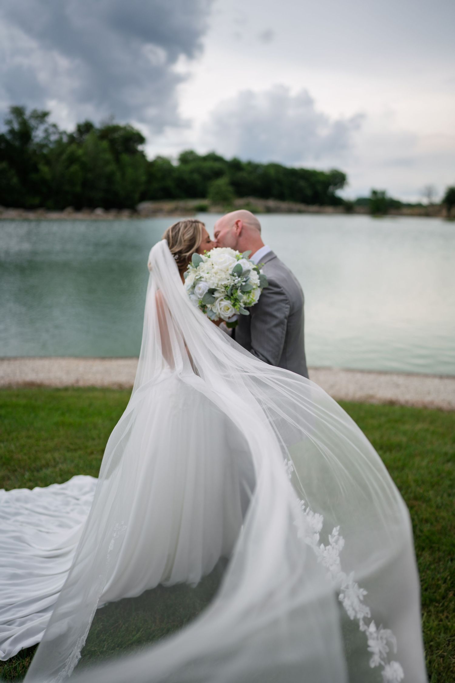 A romantic wedding scene by a lake with a bride's long white veil flowing dramatically in the wind during a cloudy day.