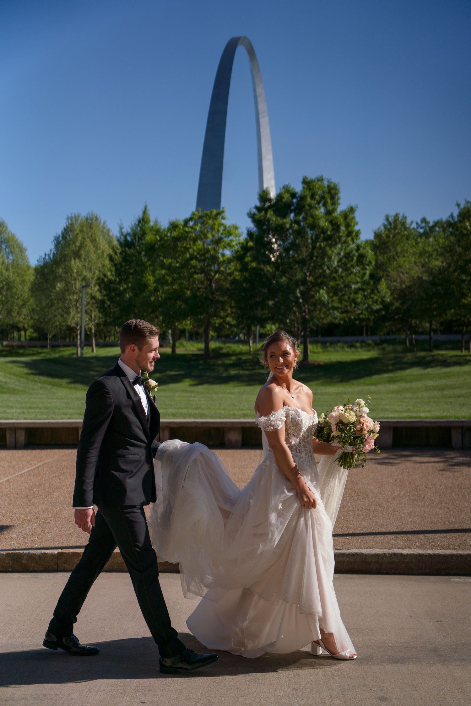 A couple in wedding attire walking together with the Gateway Arch in St. Louis visible in the background.
