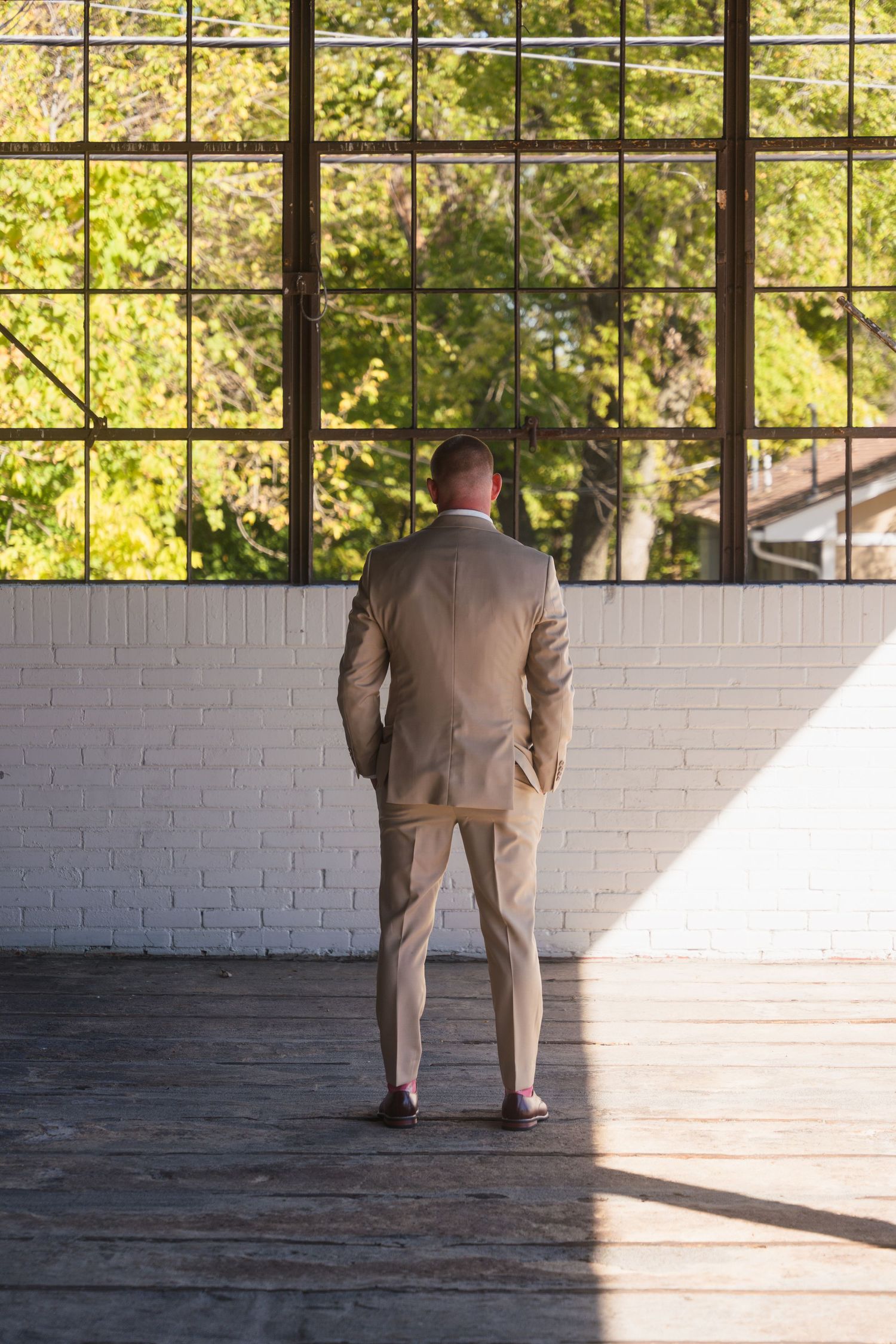 A groom in a tan suit stands silhouetted against large windows in an industrial venue.