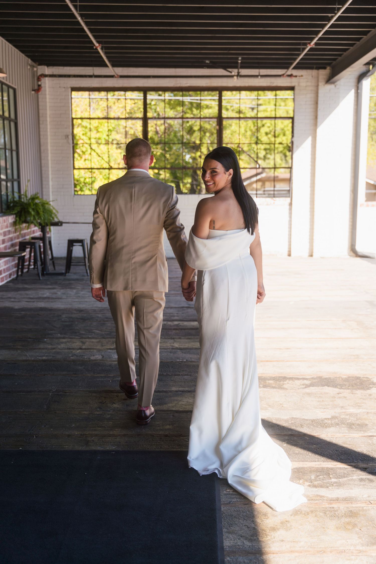 A couple in wedding attire strolls through a sunlit porch venue with large windows and a rustic interior.