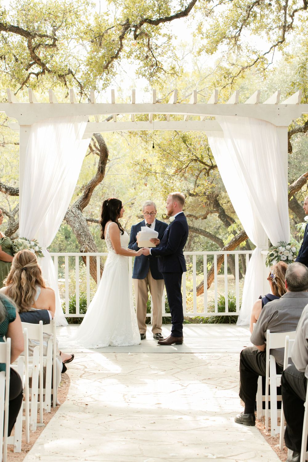 Outdoor wedding ceremony under white draped gazebo with oak trees in background.