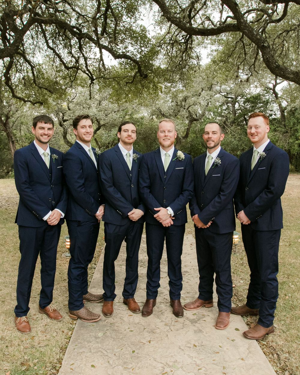 Groomsmen in navy suits and brown shoes pose under oak trees.