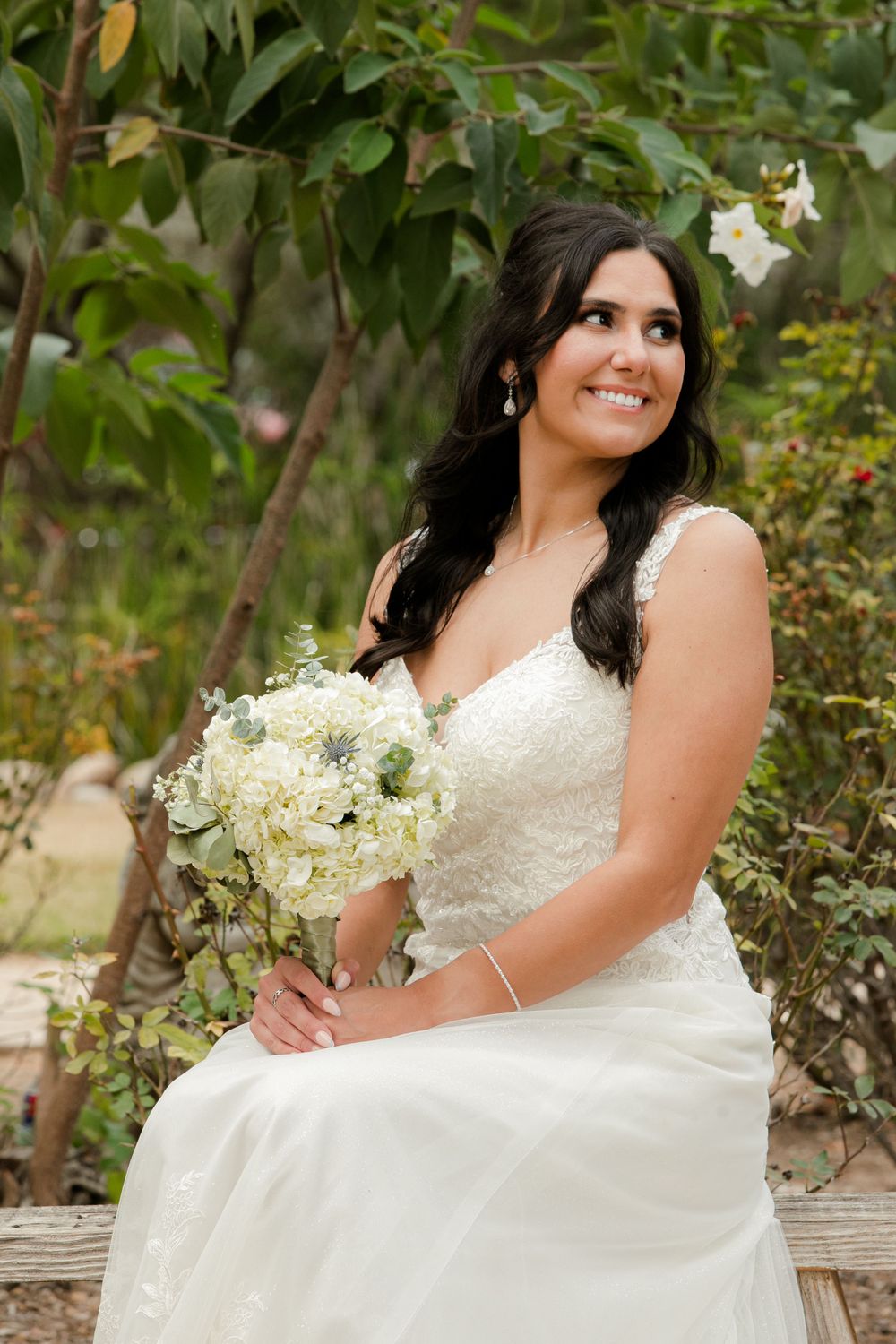 Bride in white lace dress poses with white floral bouquet in garden setting.