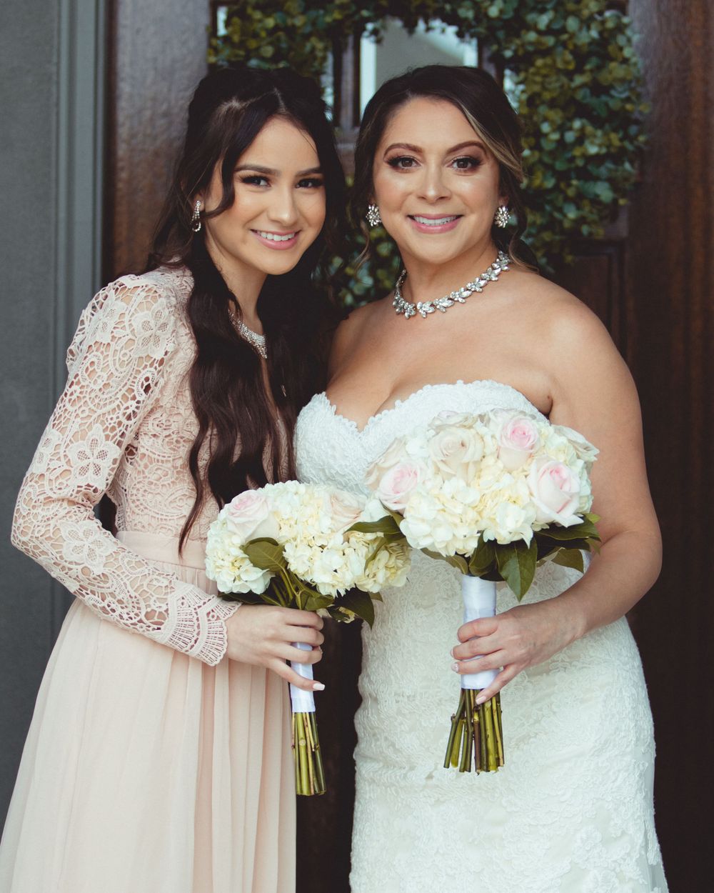 Two women in formal attire hold white floral bouquets while posing together indoors.